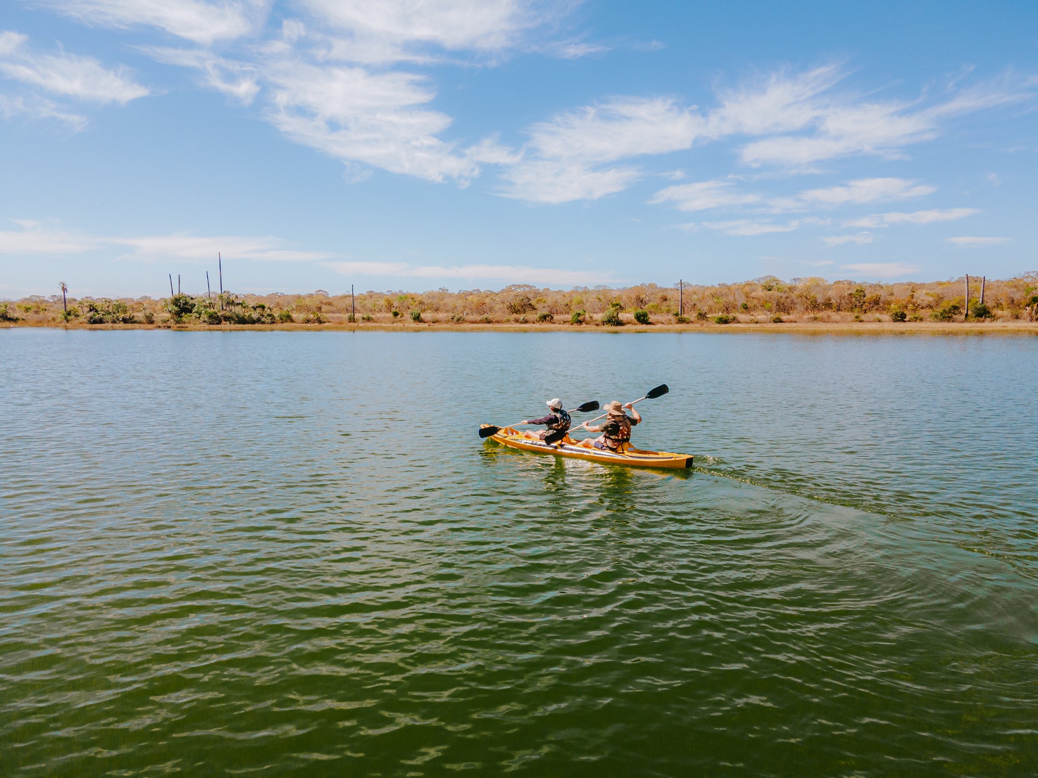 Two paddlers in a yellow tandem kayak gliding across the calm green waters of Lagoa das Araras at Pousada Trijunção, with dry Cerrado woodland stretching along the far shore beneath a sunny blue sky