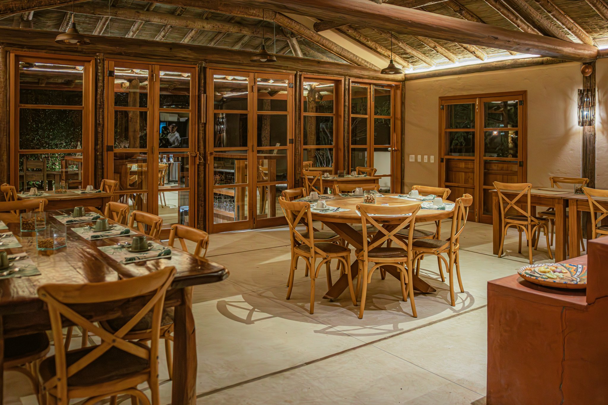 Warmly lit dining room at Pousada Trijunção featuring rustic wooden cross-back chairs, round and long communal tables set with green napkins, thatched ceiling with exposed timber beams, and large wood-framed glass doors opening to the garden