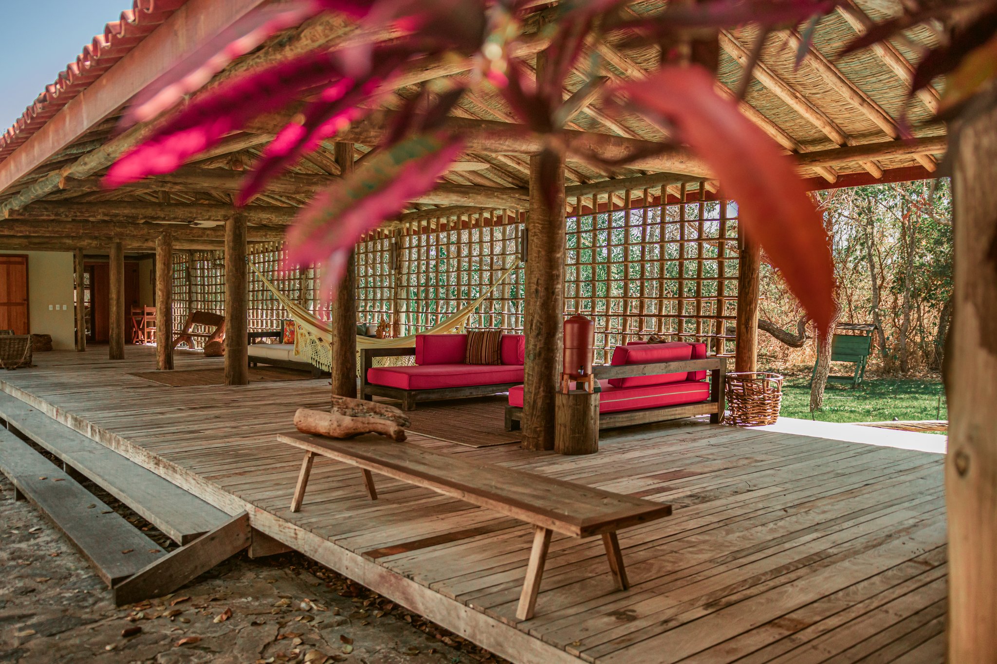 Open-air lounge at Pousada Trijunção featuring a rustic thatched roof supported by log columns, red-cushioned wooden sofas, hanging hammocks, and a wide timber deck framed by pink bougainvillea