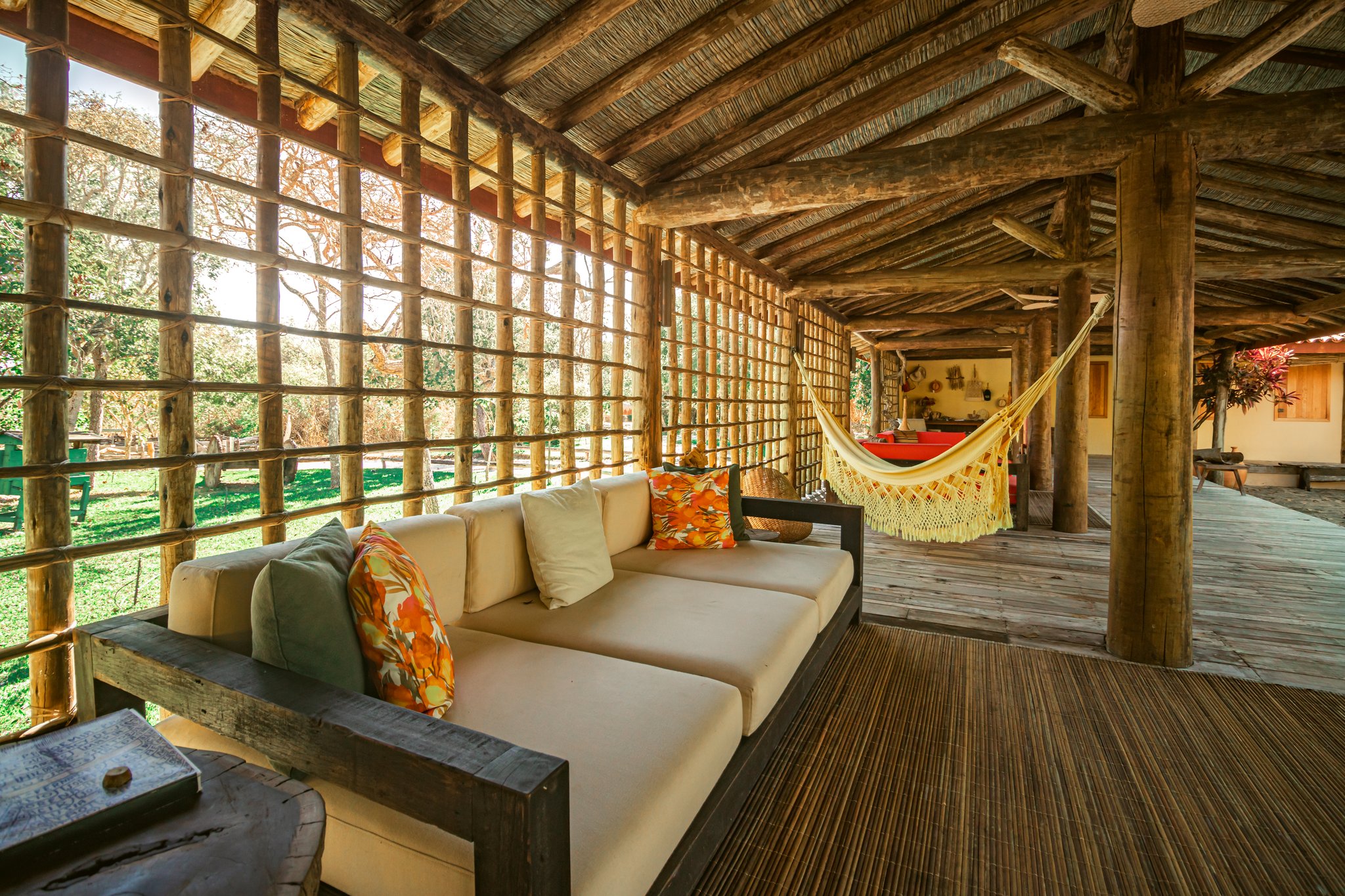 Open-air veranda at Pousada Trijunção with a cream sofa, colorful cushions, a fringed hammock, and a rustic log-and-thatch structure overlooking lush Cerrado gardens