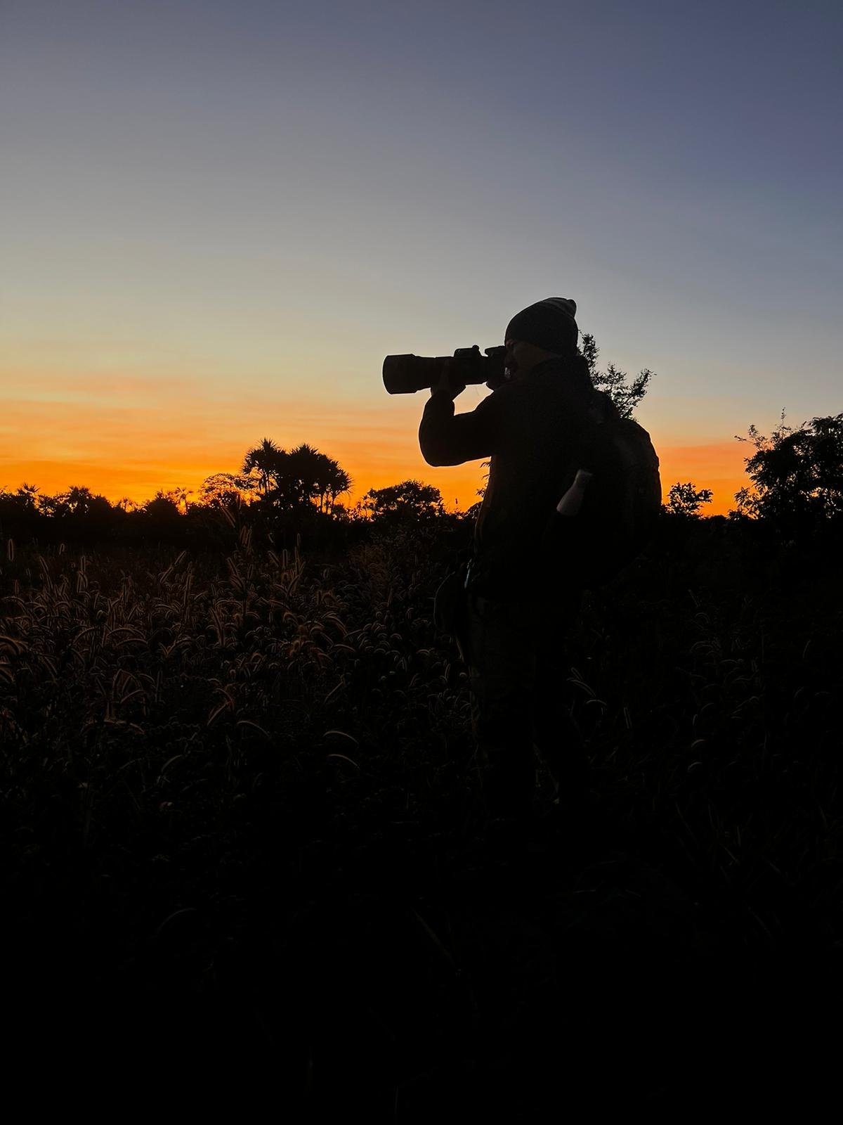 Silhouette of a wildlife photographer with a telephoto lens raised to their eye, standing among tall Cerrado grasses against a vivid orange and blue sunrise sky at Pousada Trijunção