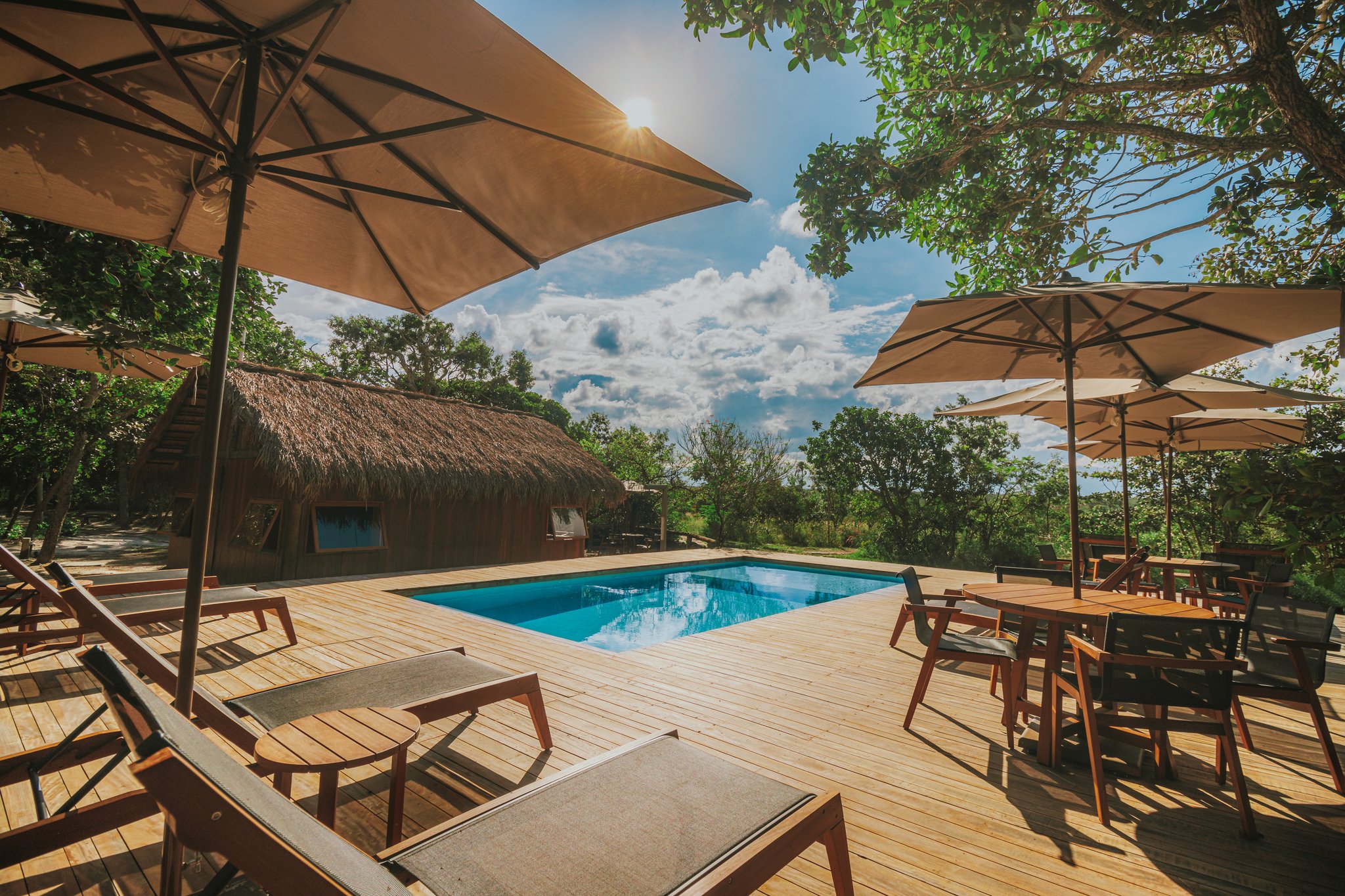 Sun-drenched wooden pool deck at Pousada Trijunção with lounge chairs, canvas umbrellas, a thatched-roof building, and green Cerrado vegetation under a blue sky
