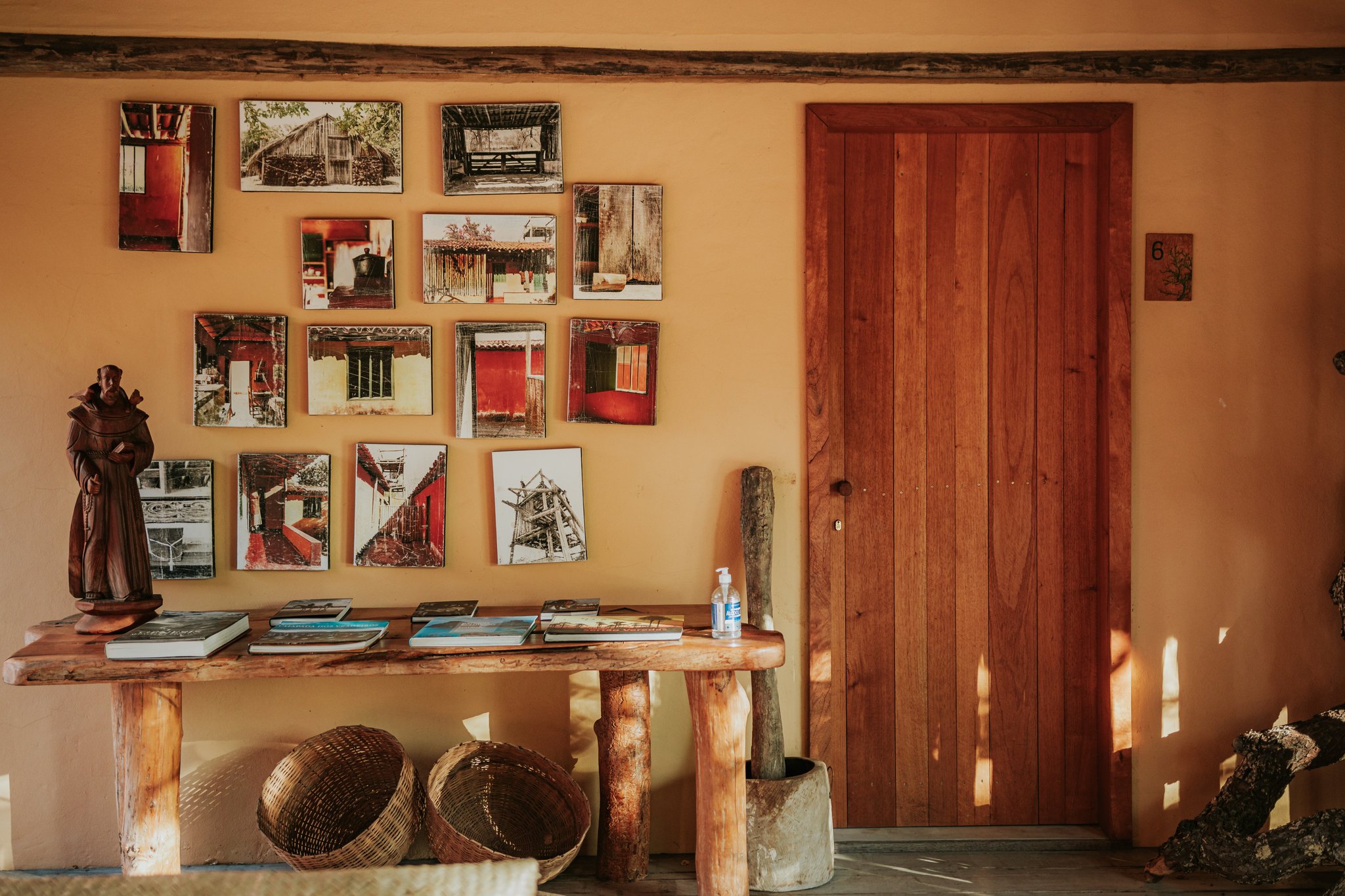 Warm ochre wall at Pousada Trijunção decorated with a grid of photographic prints depicting traditional Cerrado architecture, beside a handcrafted wooden console table holding books, a carved monk figurine and woven baskets