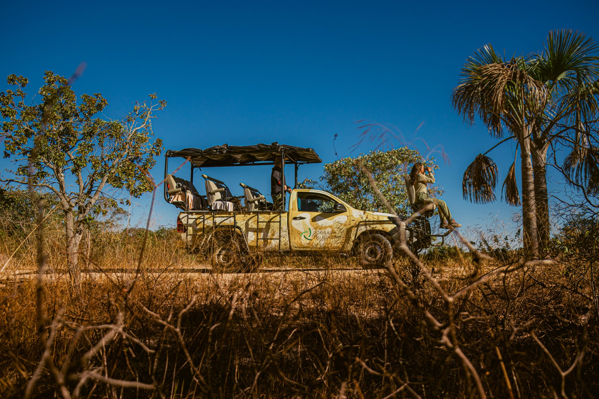 A Pousada Trijunção open safari vehicle parked on a dirt track in the dry Cerrado savanna, with a guest perched on the side rack using binoculars beneath a deep blue sky