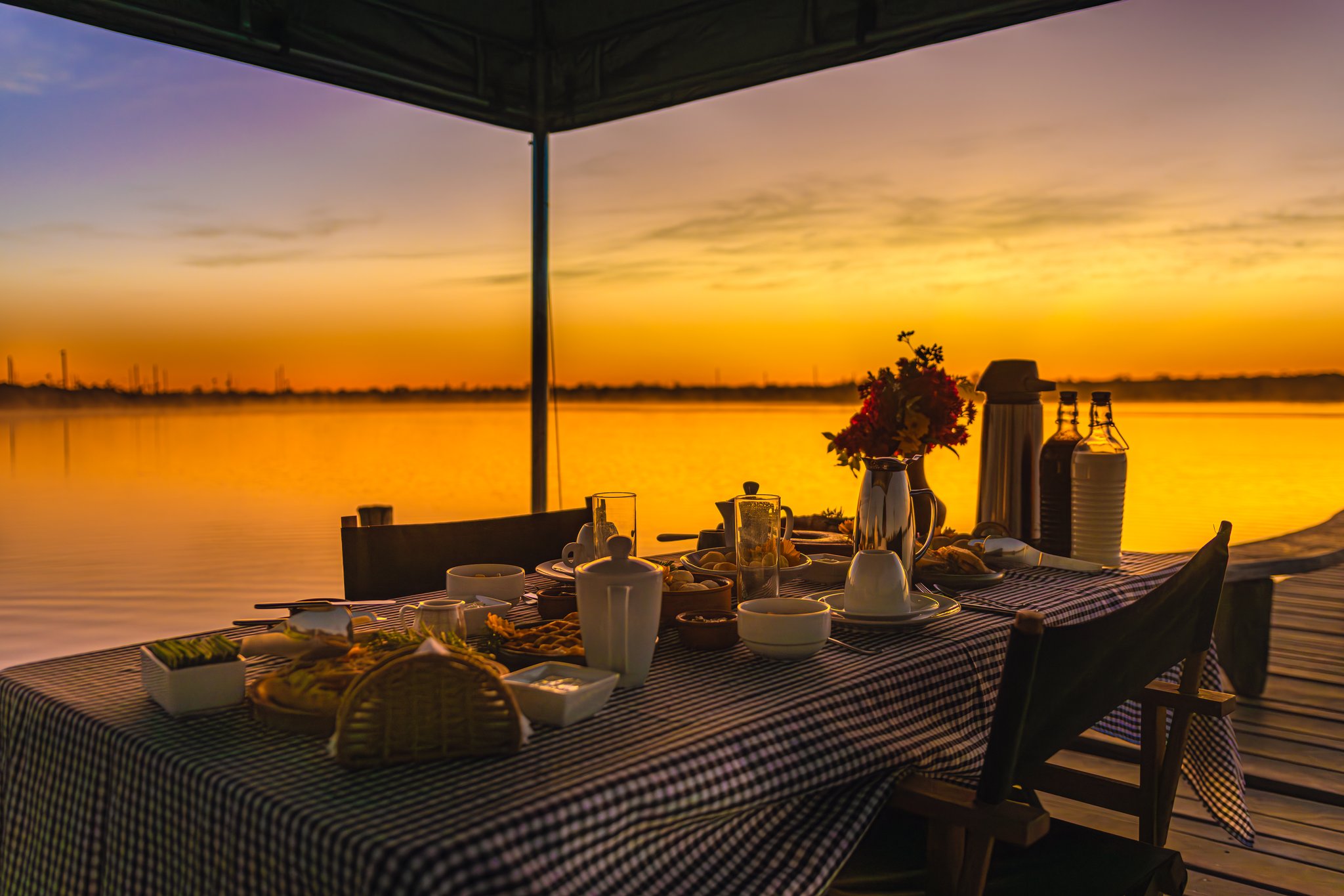 A fully set breakfast table on a wooden pier at Pousada Trijunção, with a gingham tablecloth, fresh flowers, coffee pots and fruit, silhouetted against a vivid golden sunrise over a glassy lake