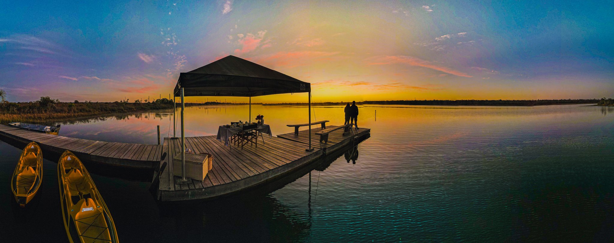 A couple silhouetted at the end of a wooden pier at Pousada Trijunção, a canopied breakfast table behind them and kayaks moored to the left, as a golden sunrise blazes across the glassy lake