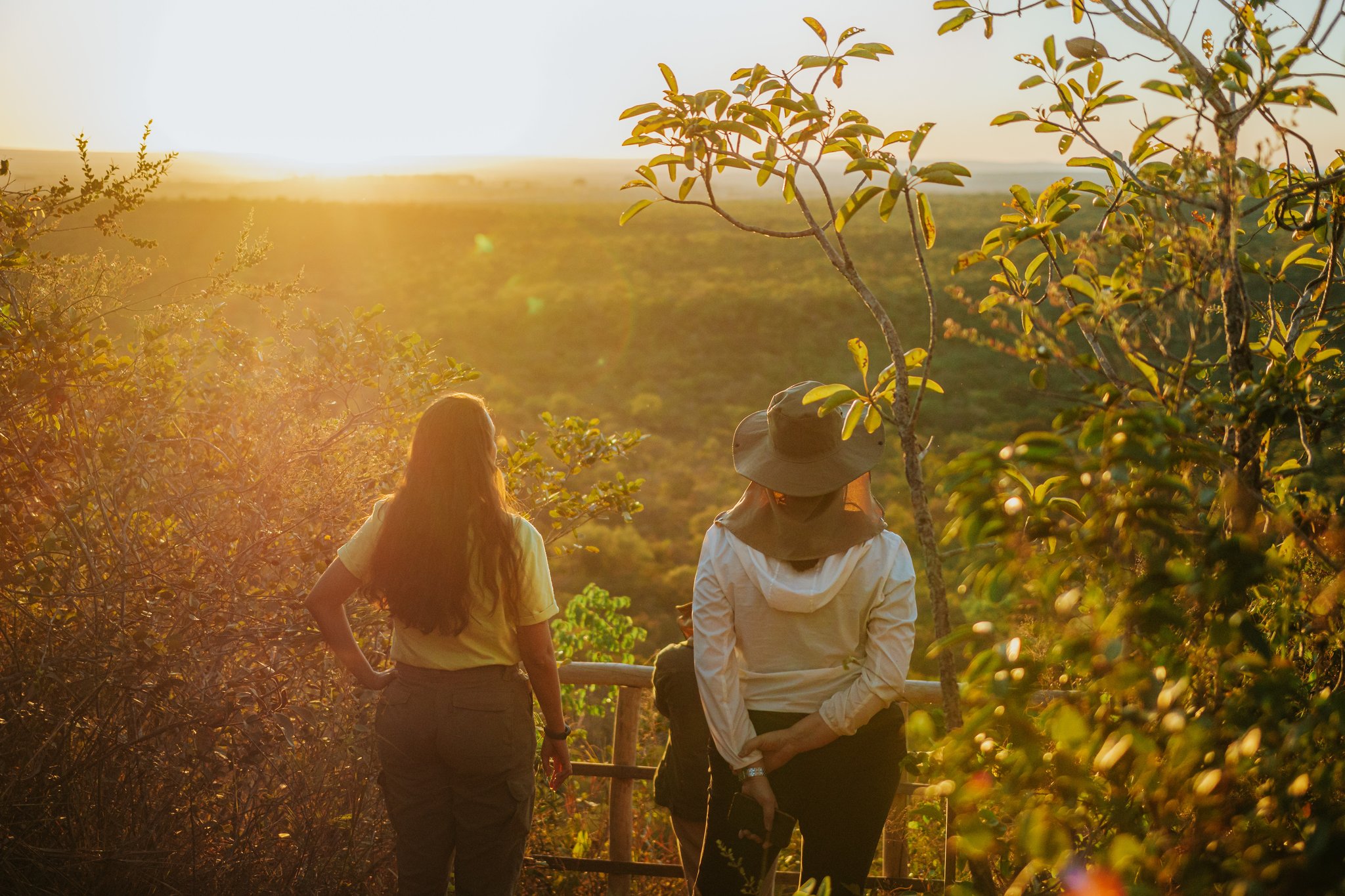 Two women standing at a wooden railing viewpoint at Pousada Trijunção, gazing over a vast golden-lit Cerrado landscape at sunset