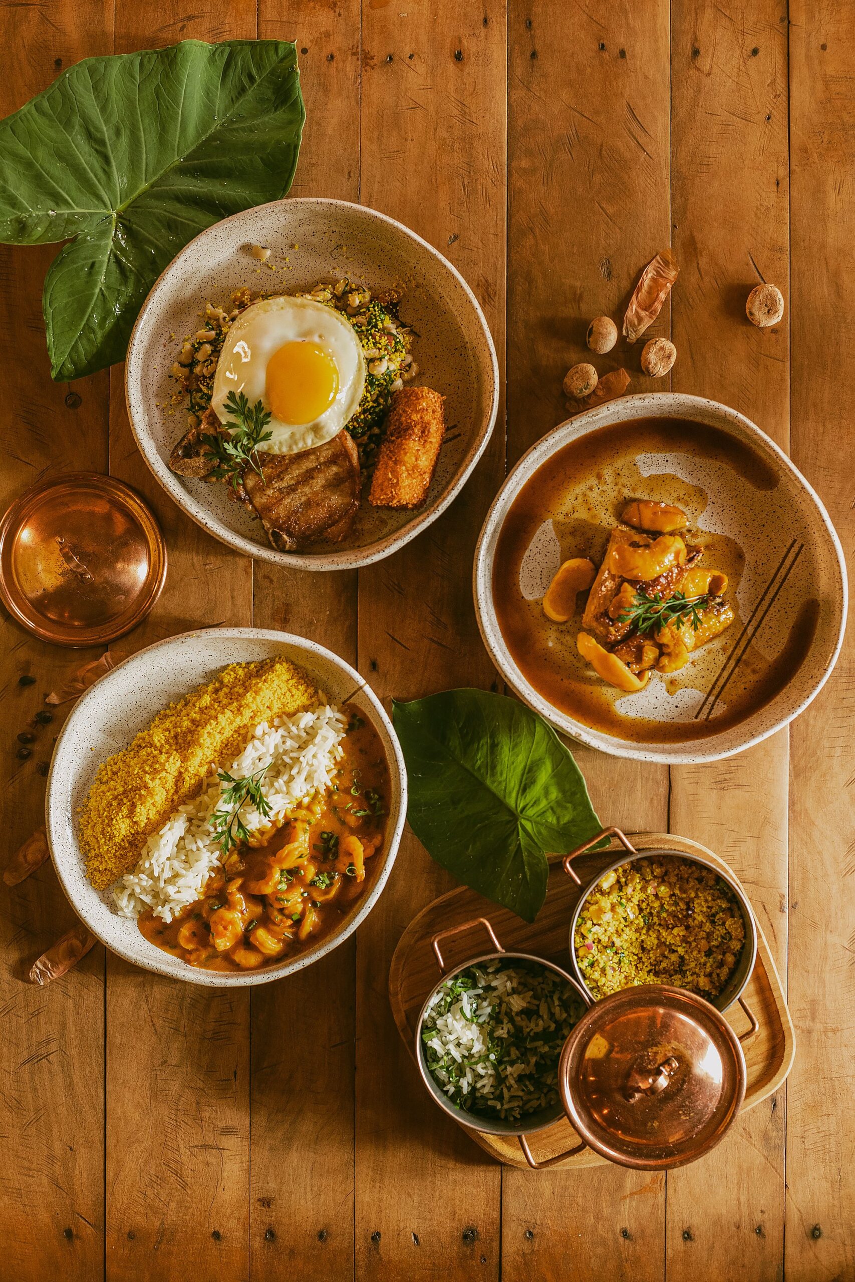 Four ceramic bowls of traditional Brazilian dishes served at Pousada Trijunção, including shrimp moqueca with rice and farofa, a fried-egg rice bowl with crispy pork belly, a rich meat stew, and copper pots of sides on a warm wooden table