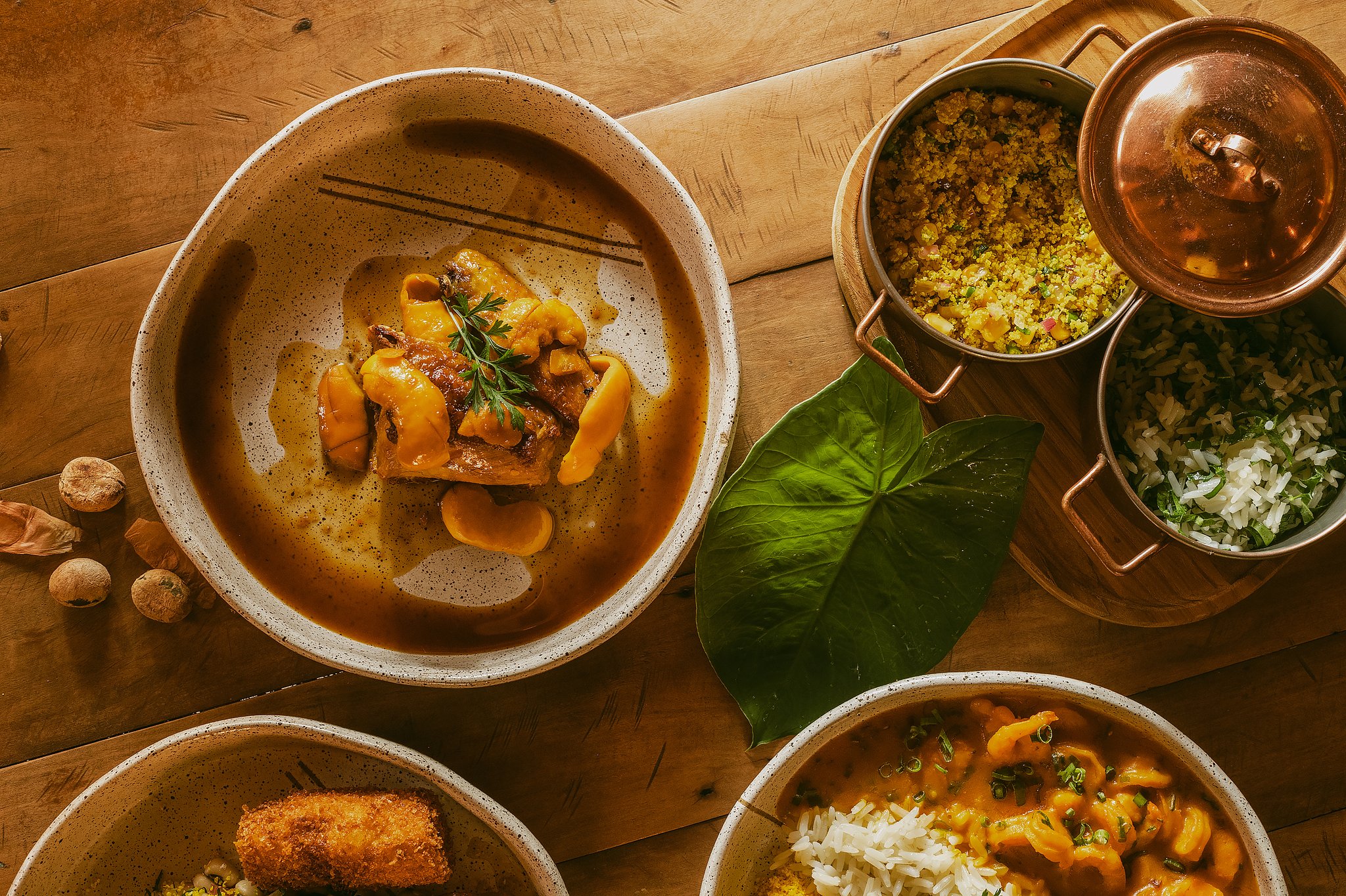 Overhead view of a Pousada Trijunção meal spread featuring a rich braised meat stew in a speckled ceramic bowl, golden farofa and herb rice in copper pots, and a creamy seafood stew with white rice, all arranged on a rustic wooden table with a tropical leaf