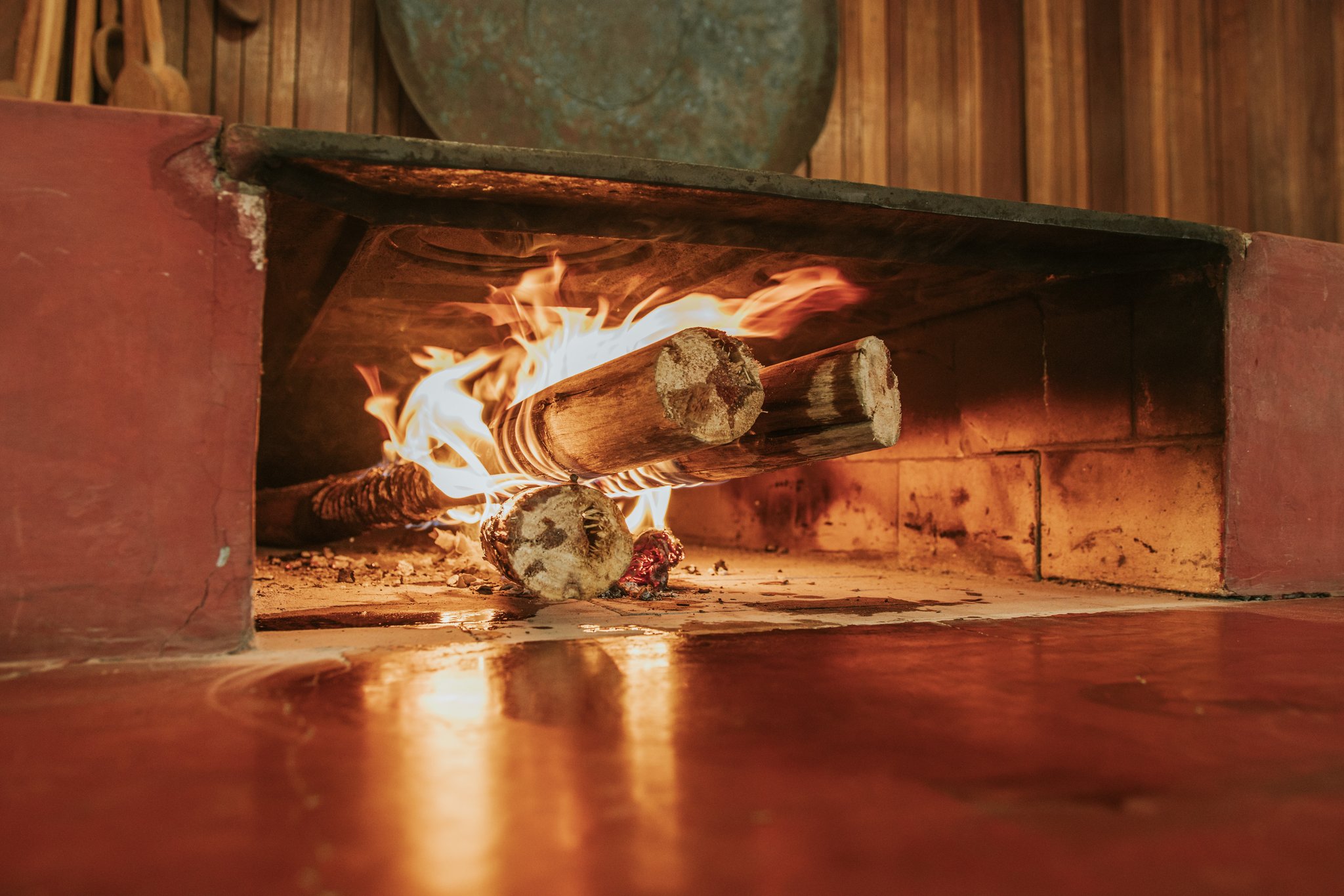Thick wooden logs burning with open flames inside a traditional wood-fired stone hearth at Pousada Trijunção, with warm firelight reflecting off the polished red floor