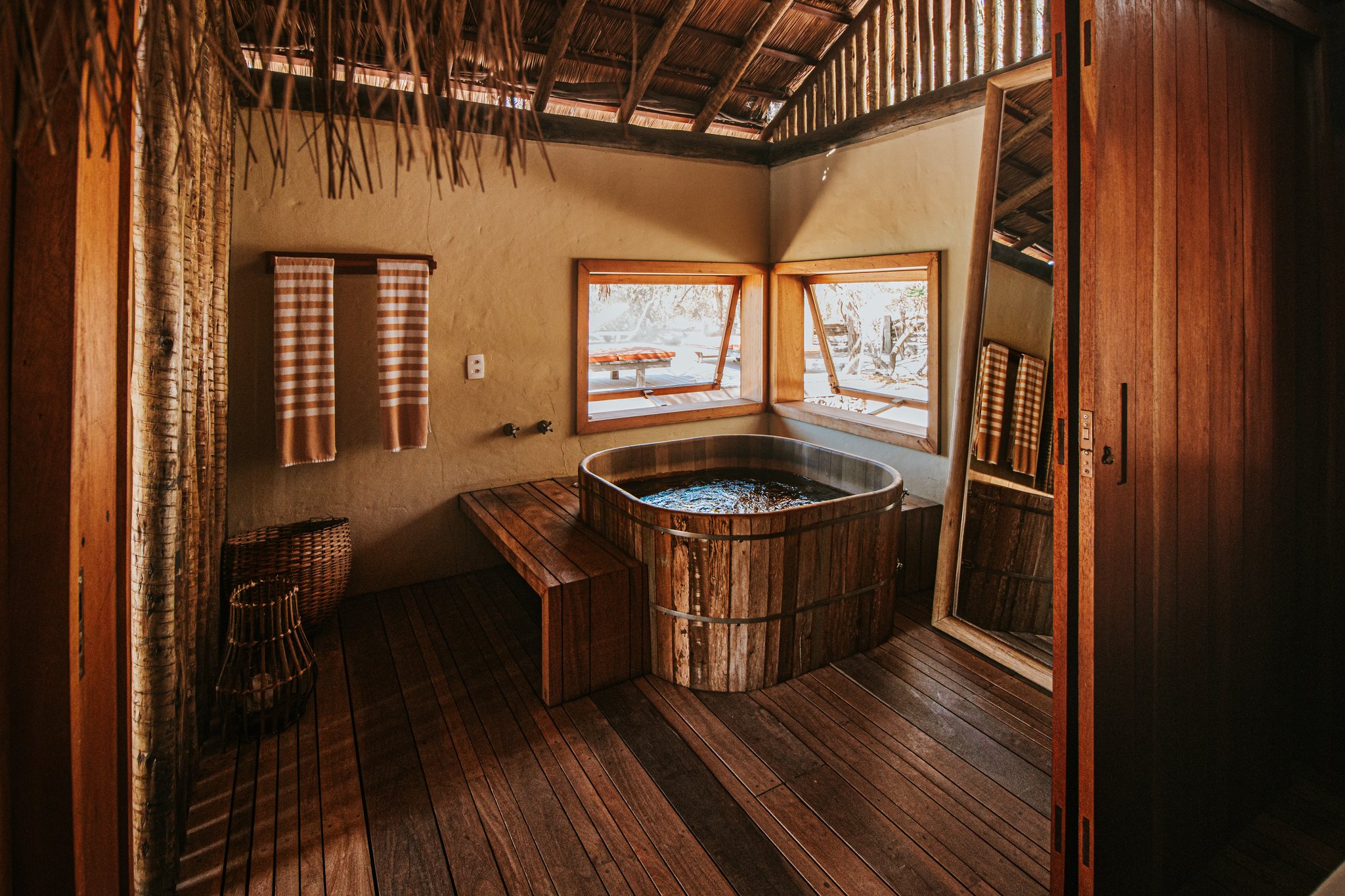 Rustic wooden barrel bathtub inside a Pousada Trijunção suite with thatched ceiling, warm timber floors and open windows overlooking the Cerrado landscape