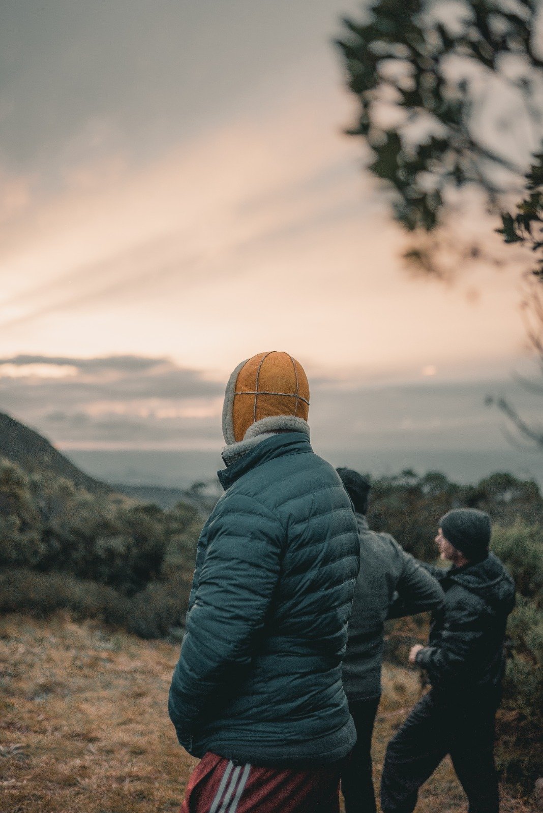 Two hikers in heavy winter layers and a sheepskin hat stand on an exposed ridge during Serra Fina Trekking, gazing over misty mountain valleys beneath a soft pastel sunrise sky in the Serra da Mantiqueira