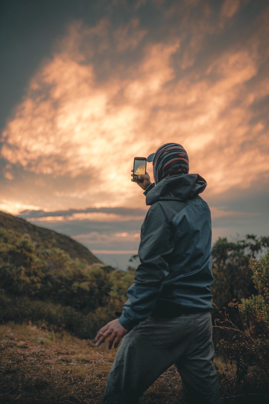 A hiker in a jacket and striped beanie photographs a dramatic orange and gold sunset sky above the Serra da Mantiqueira ridgeline during a Serra Fina Trekking expedition