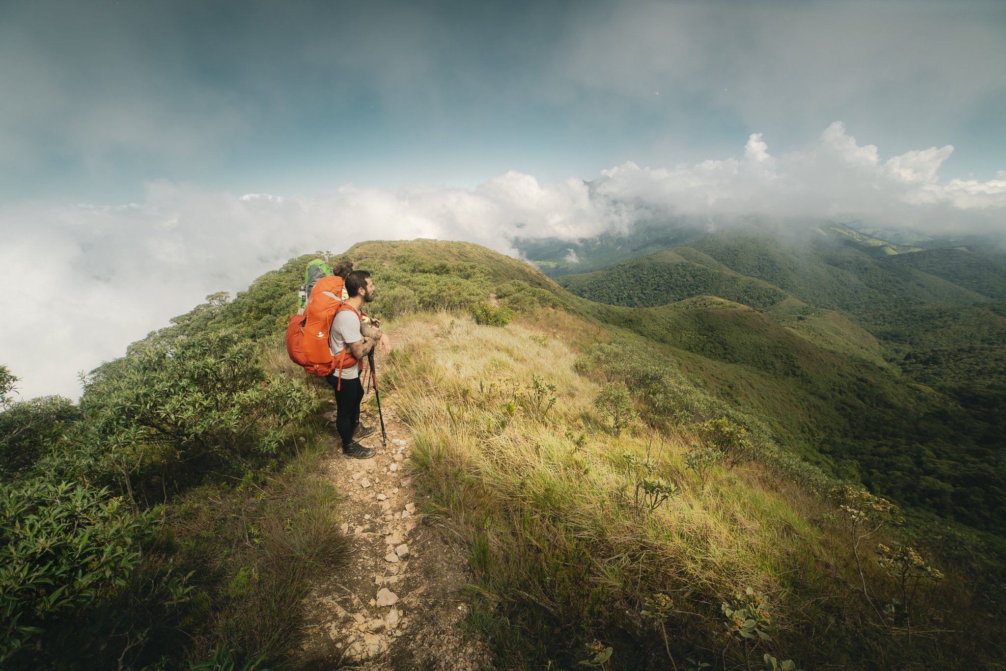 A hiker with an orange backpack and trekking poles pauses on a narrow exposed ridge during a Serra Fina Trekking traverse, with rolling cloud-covered peaks of the Serra da Mantiqueira stretching to the horizon