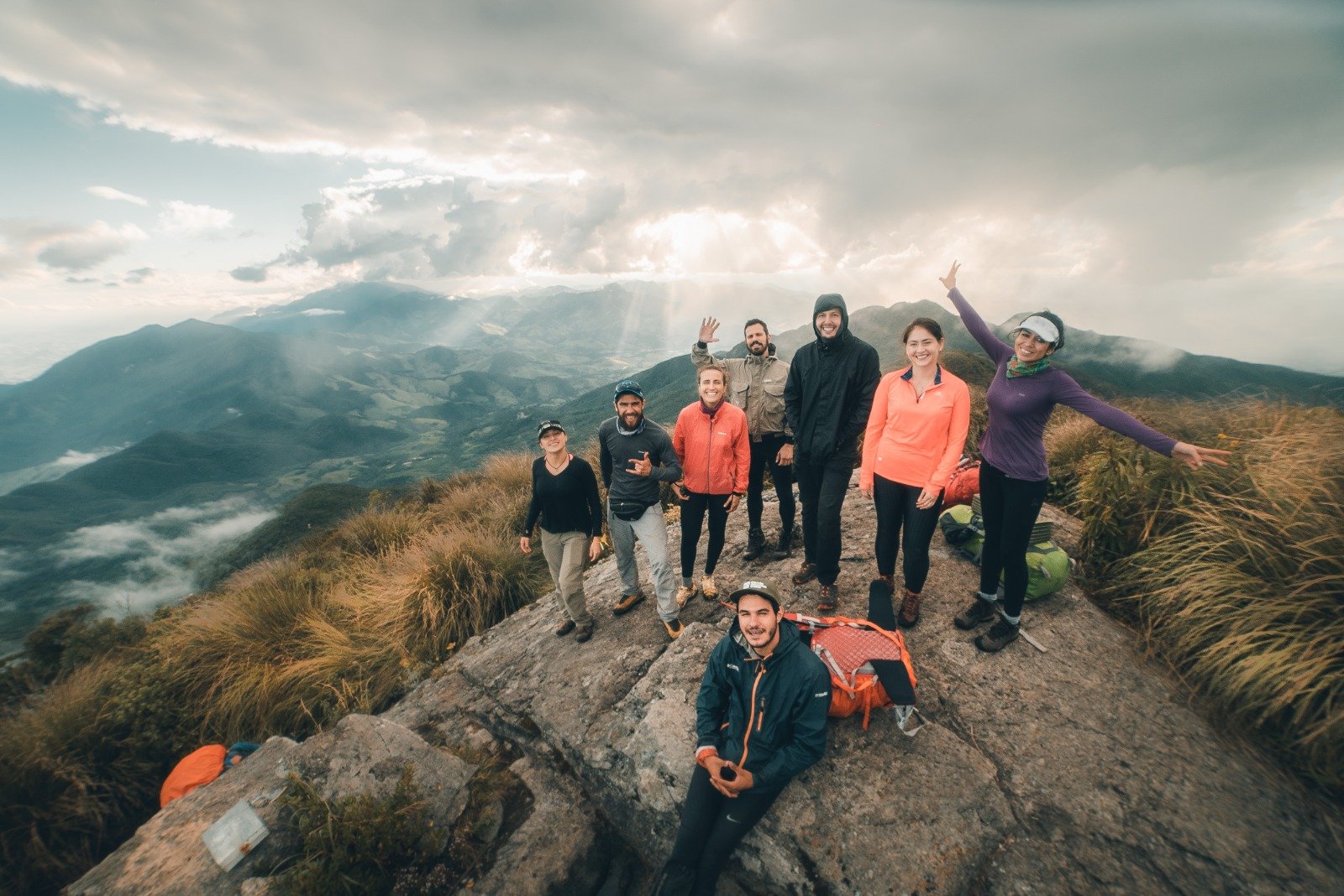 Eight hikers celebrate on a rocky summit during the Serra Fina Trekking, with dramatic cloud-filled skies and the rolling ridges of the Serra da Mantiqueira stretching to the horizon