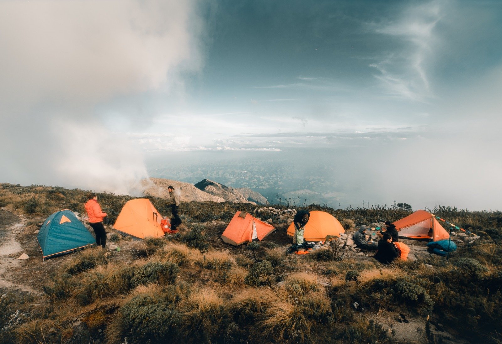 A group of trekkers setting up orange and teal tents at a Serra Fina Trekking high-altitude wild camp on an exposed ridge in the Serra da Mantiqueira, with sweeping views of valleys and dramatic storm clouds above