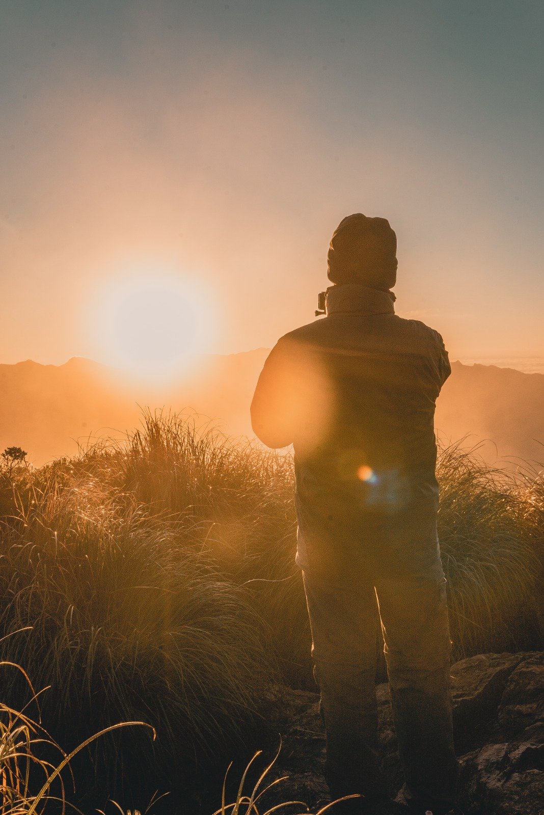 A hiker's silhouette stands among golden mountain grasses at sunrise during a Serra Fina Trekking expedition, with misty ridges of the Serra da Mantiqueira glowing orange in the background