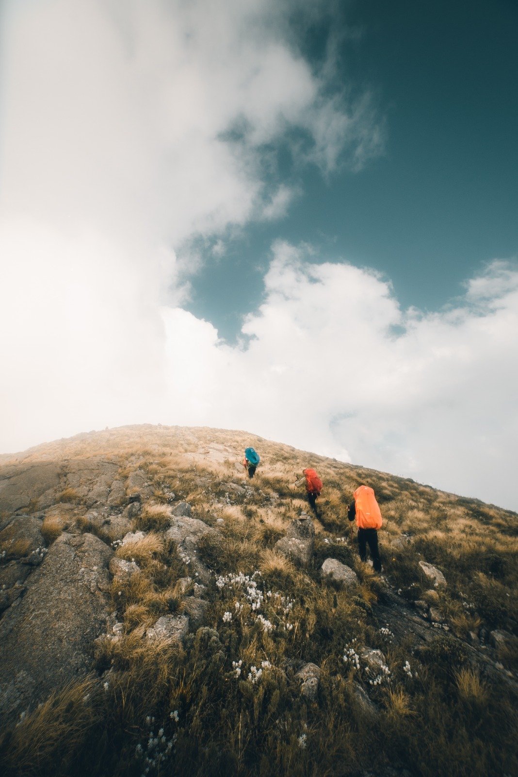 Three hikers with colourful backpacks ascending a steep rocky ridge on the Serra Fina Trekking route in the Serra da Mantiqueira, under a dramatic sky