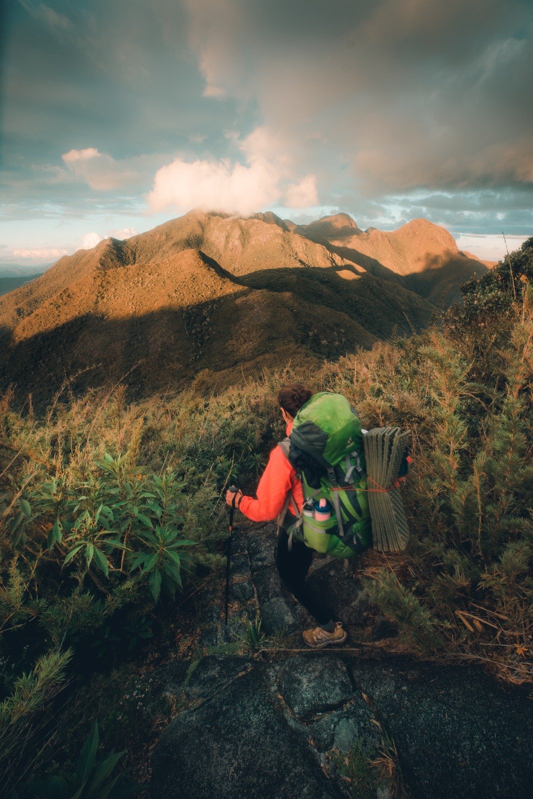 A backpacker in an orange jacket carrying a full pack pauses on a rocky ridge during Serra Fina Trekking, with golden-lit mountain summits of the Serra da Mantiqueira stretching under a dramatic cloudy sky