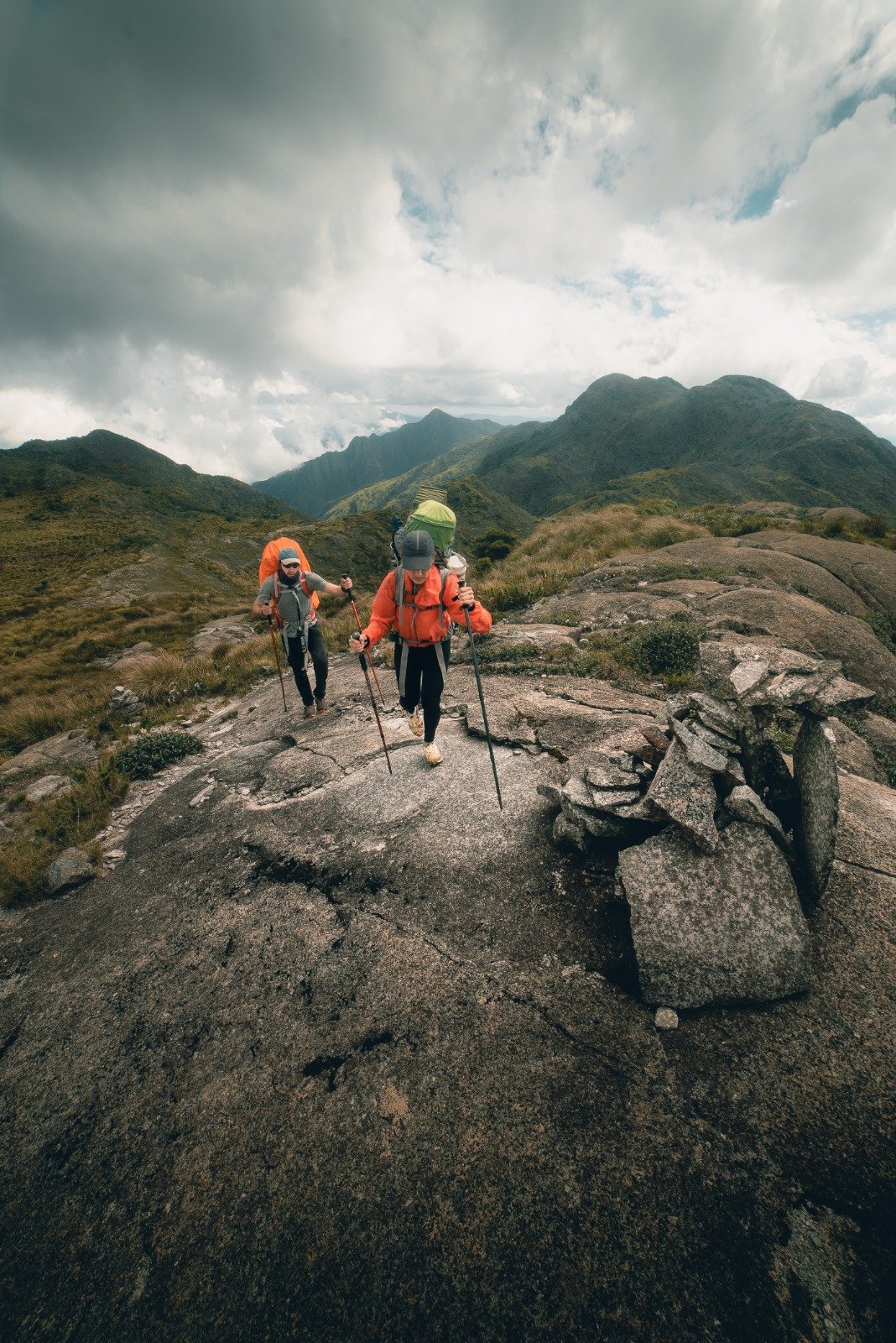 Two backpackers in orange jackets using trekking poles to navigate a granite rock slab on the Serra Fina Trekking route, with green mountain ridges and dramatic clouds filling the Serra da Mantiqueira skyline behind them