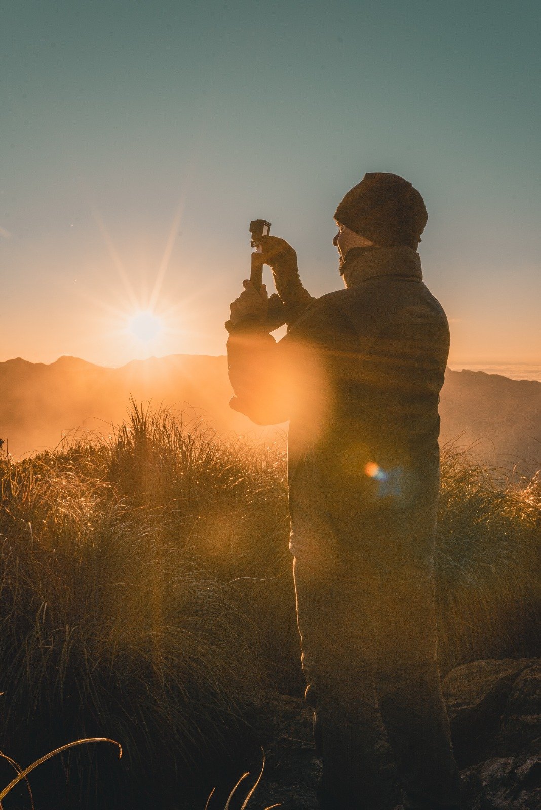 A hiker in a beanie and puffer jacket photographs the golden sunrise over misty Serra da Mantiqueira ridges during a Serra Fina trekking expedition, wild grasses glowing in the foreground