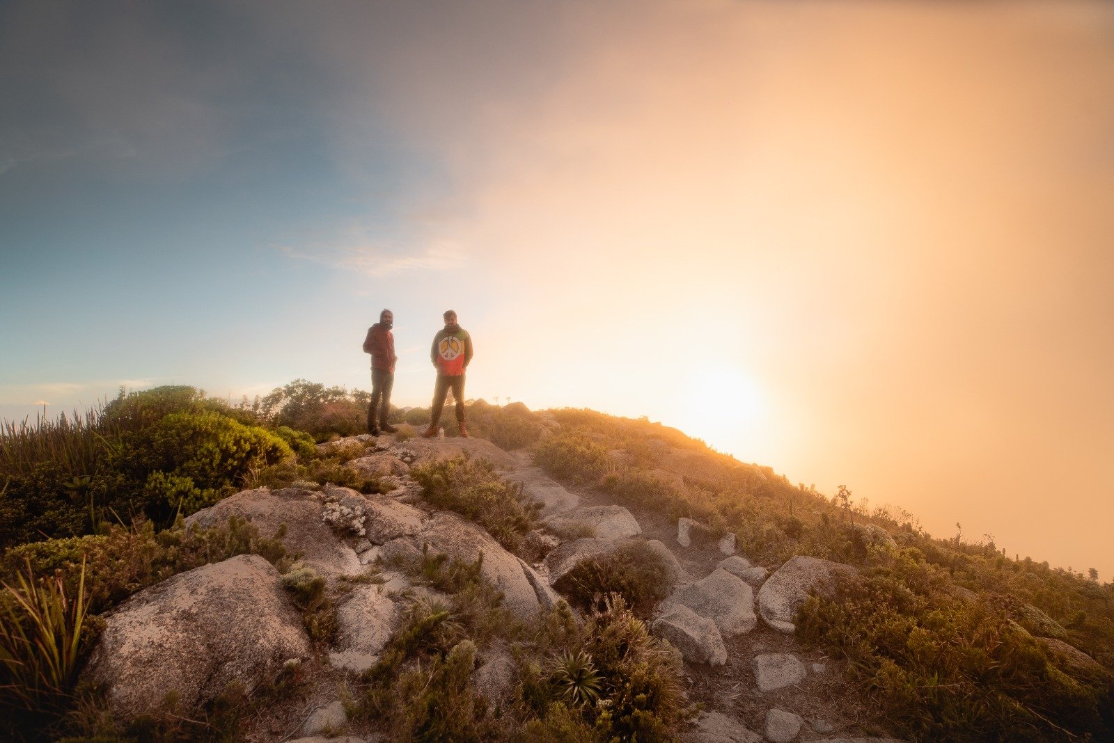 Two hikers silhouetted against a glowing golden sunrise on a rocky Serra Fina Trekking summit, surrounded by high-altitude Atlantic forest vegetation in the Serra da Mantiqueira.