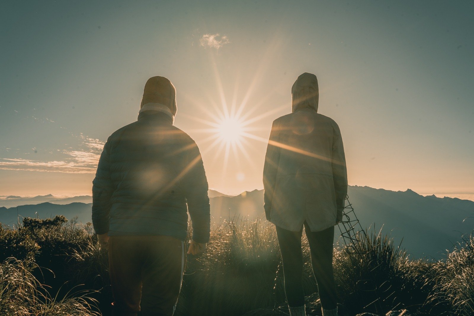 Two hooded hikers silhouetted against a brilliant starburst sunrise on a Serra Fina Trekking summit, with layers of Mantiqueira mountain ridges stretching to the horizon