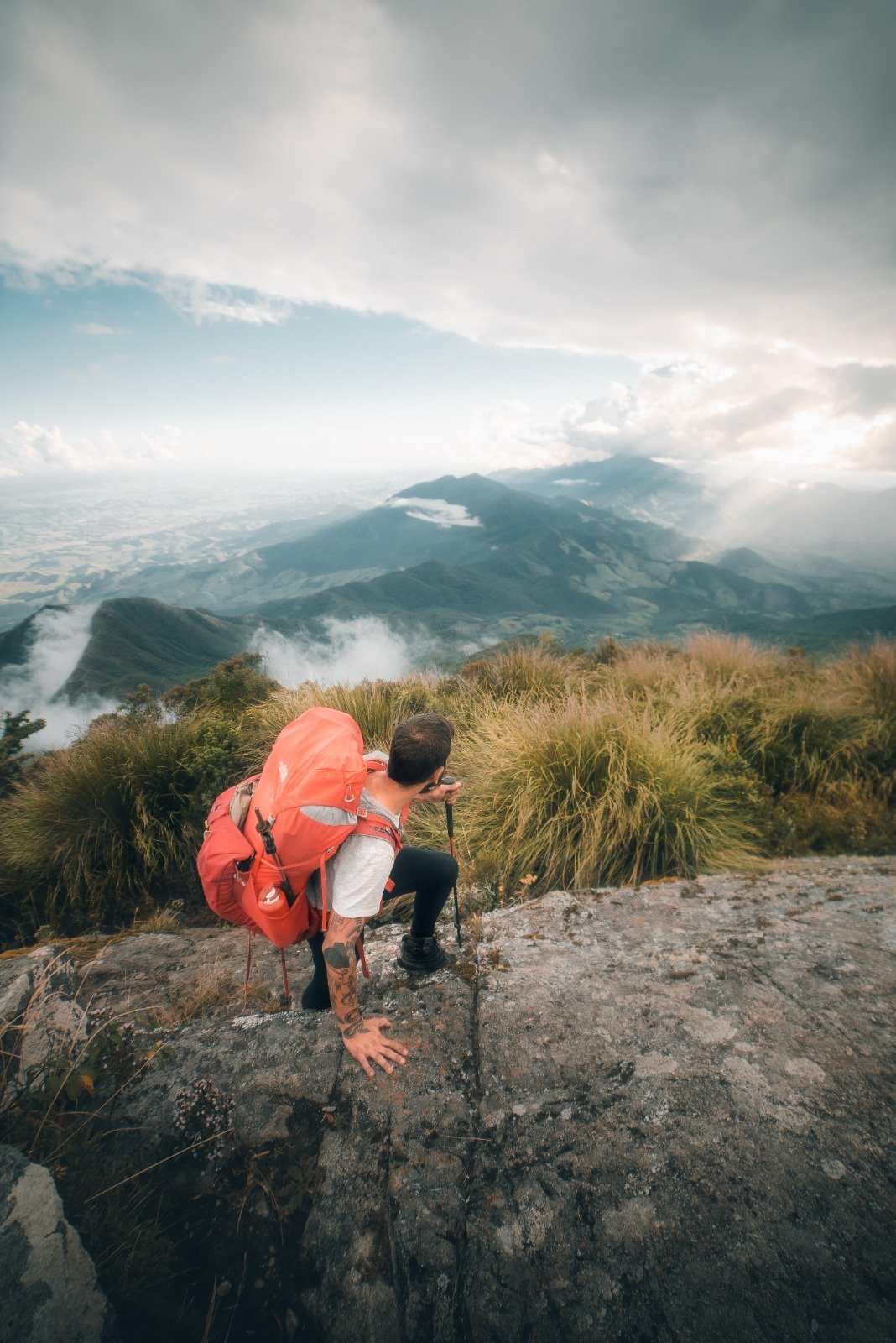 A hiker with a large red backpack and trekking pole crouches on a rocky ledge during Serra Fina Trekking, gazing over a vast misty mountain landscape in the Serra da Mantiqueira