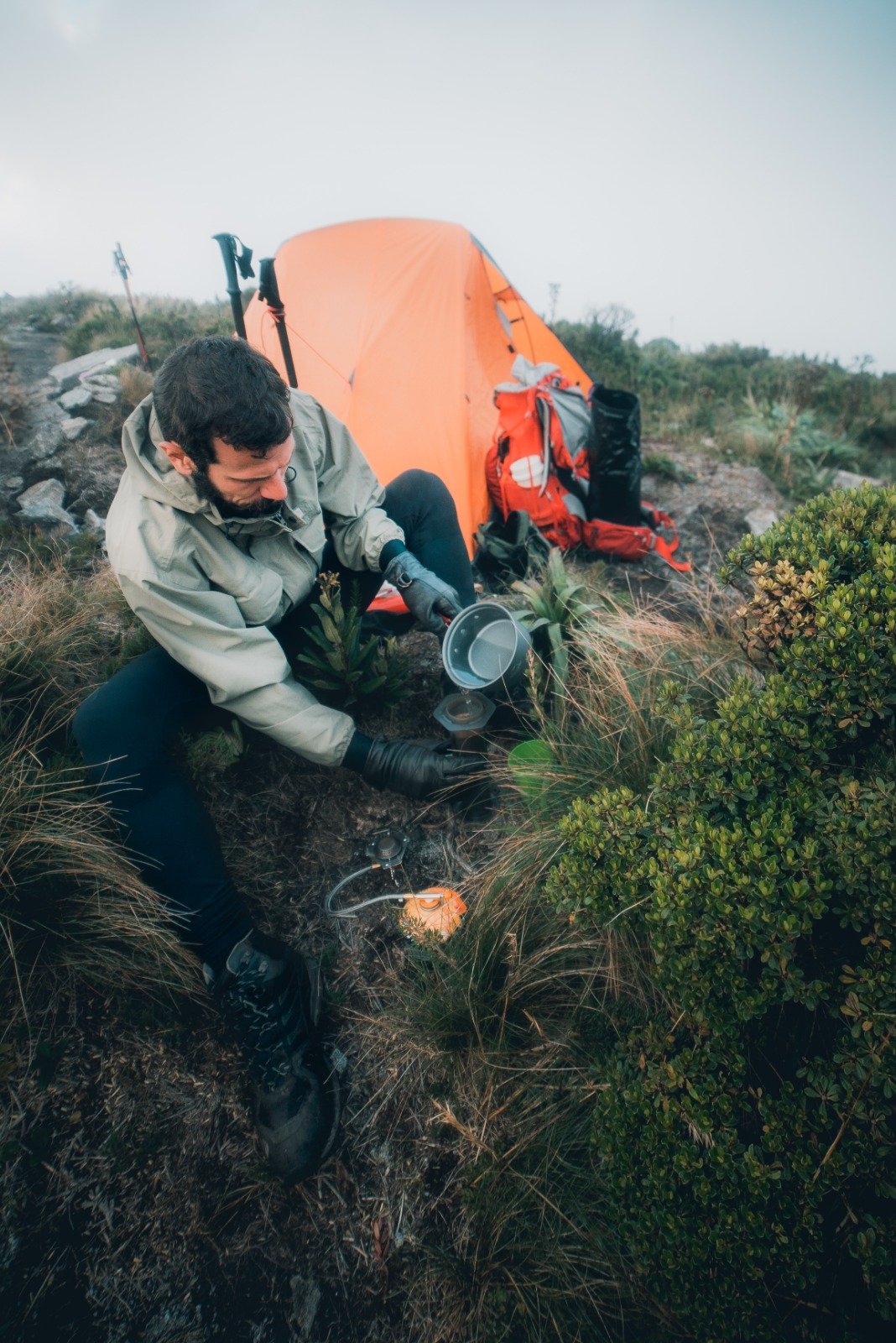 A bearded hiker in a rain jacket pours water into a pot on a compact camp stove beside an orange tent and red backpack during a Serra Fina Trekking expedition in the Serra da Mantiqueira highlands