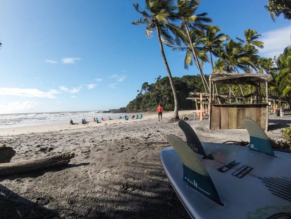 A surfboard with FCS fins rests on the sand in the foreground while a group takes surf lessons in Itacaré on a palm-fringed Atlantic beach, with a rustic bamboo hut and lush rainforest backdrop under a clear blue sky