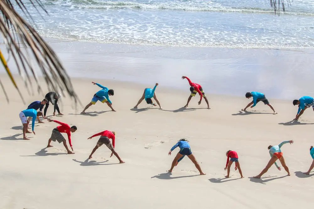A large group of surf students doing synchronised warm-up stretches on a wide sandy beach in Itacaré during surf lessons, with gentle Atlantic waves in the background