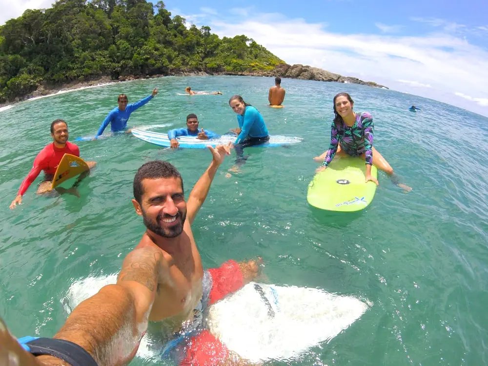 A group of smiling students holding colorful surfboards in the turquoise waters of Itacaré during surf lessons in Itacaré, with a lush forested headland in the background