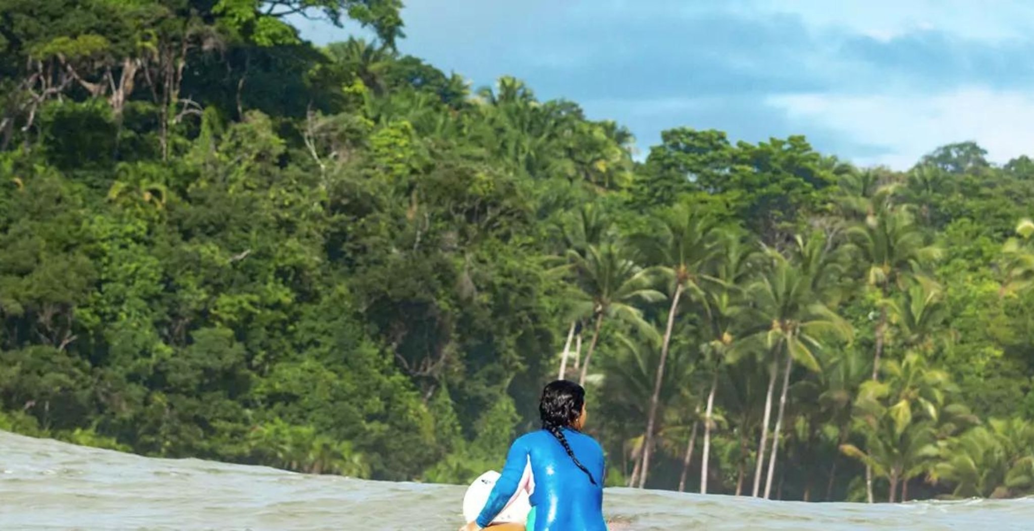 A surfer in a blue wetsuit carrying a board wades into the ocean in front of a dense tropical rainforest on the coast of Itacaré, Bahia