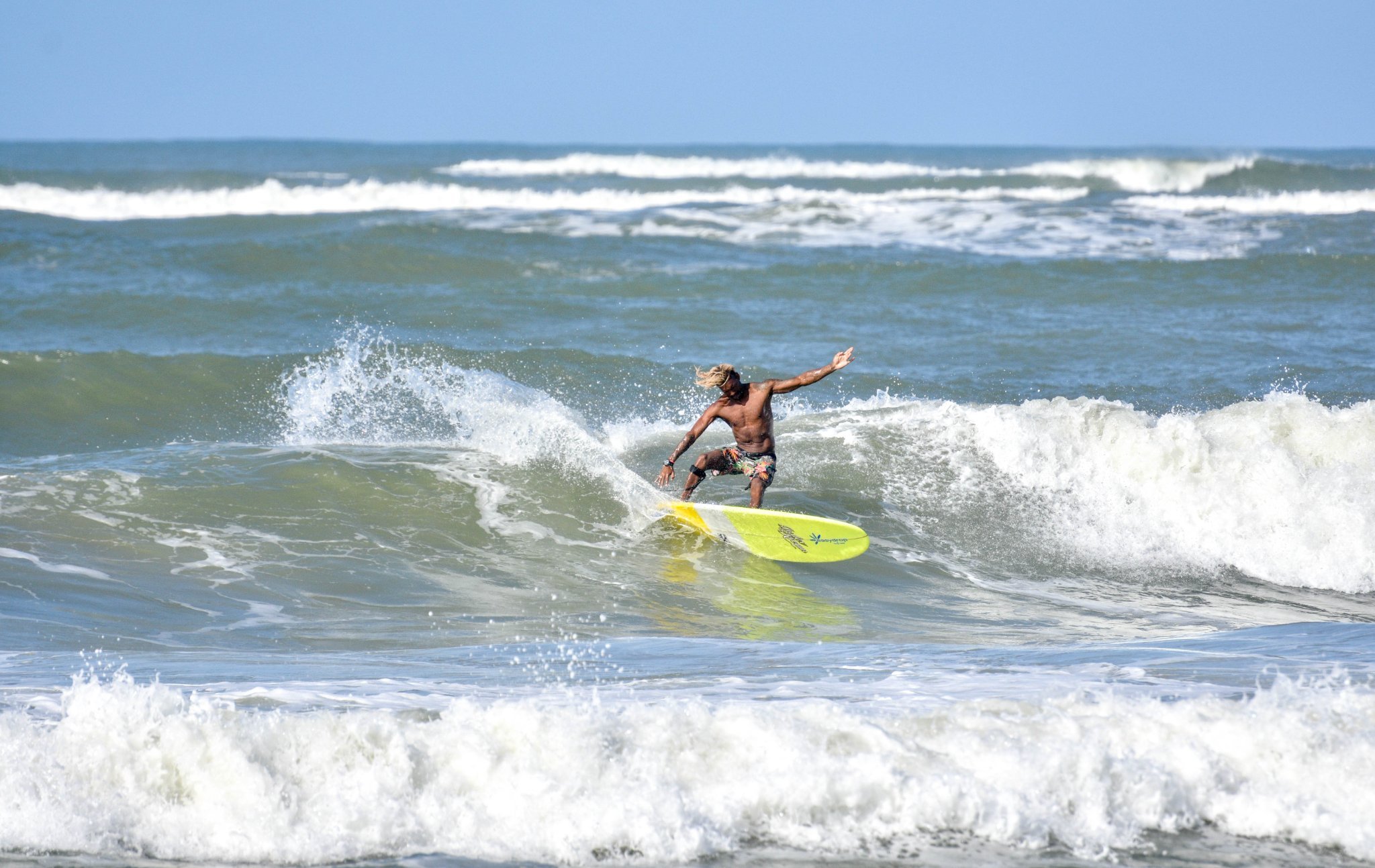 A surfer rides a breaking wave on a yellow longboard during surf lessons in Itacaré, arms outstretched for balance under a clear blue sky