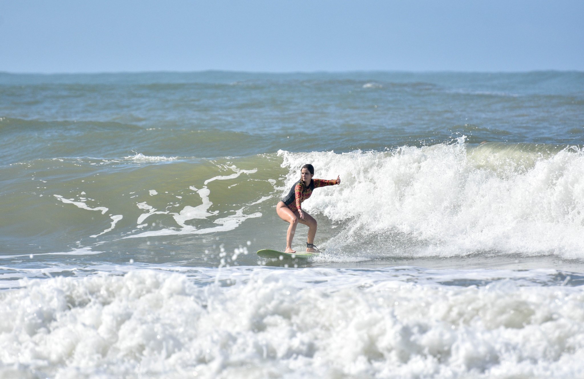 A woman in a floral rash guard crouches low on a surfboard, riding a foaming wave at a surf school in Itacaré, Bahia