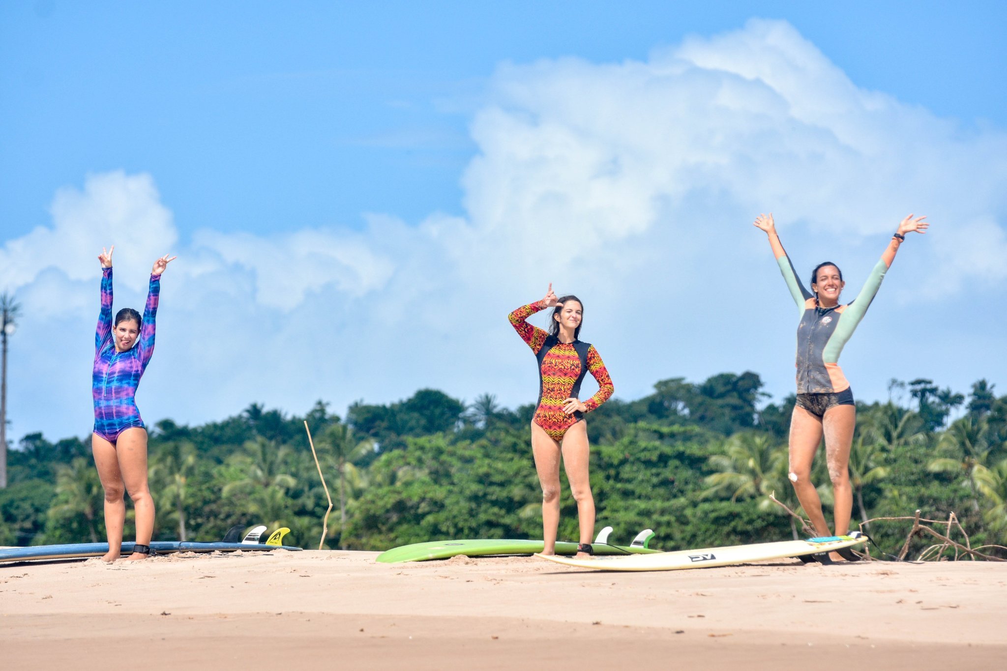 Three smiling women in colourful rash guards raise their arms in celebration on a sandy beach at this surf school in Itacaré, with surfboards on the sand and lush tropical forest under a blue sky behind them