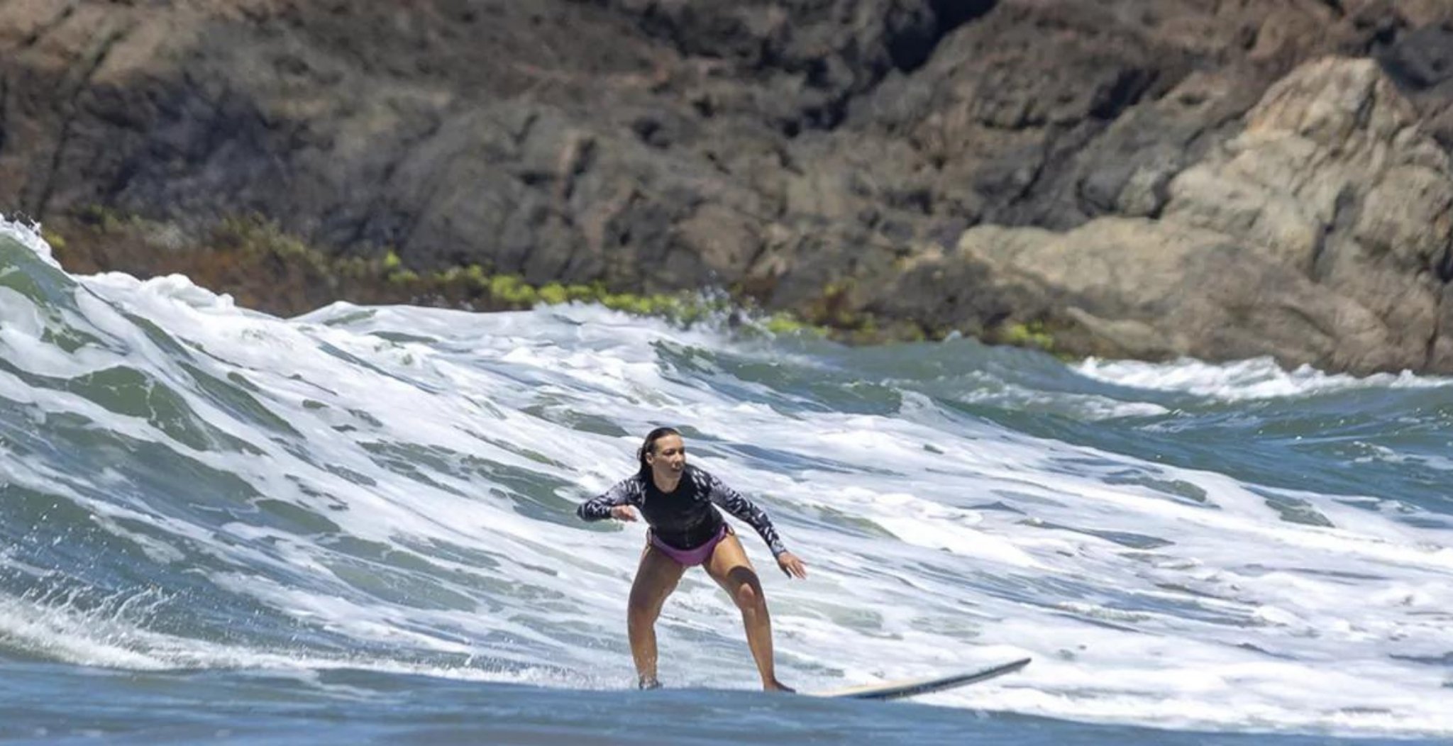 A woman in a long-sleeve rash guard rides a foaming wave at a surf school in Itacaré, with dramatic dark rocky cliffs rising in the background
