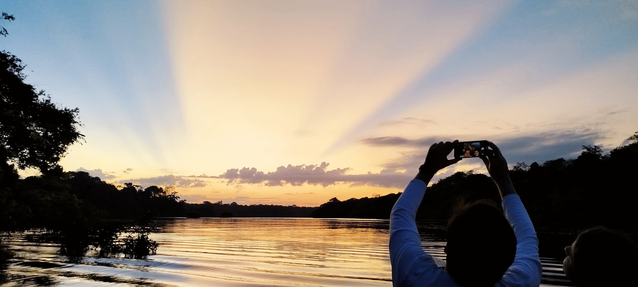 Crepuscular rays fan across the sky as a traveler captures the moment. Traveler photographing dramatic sunrays over the Amazon river at sunset