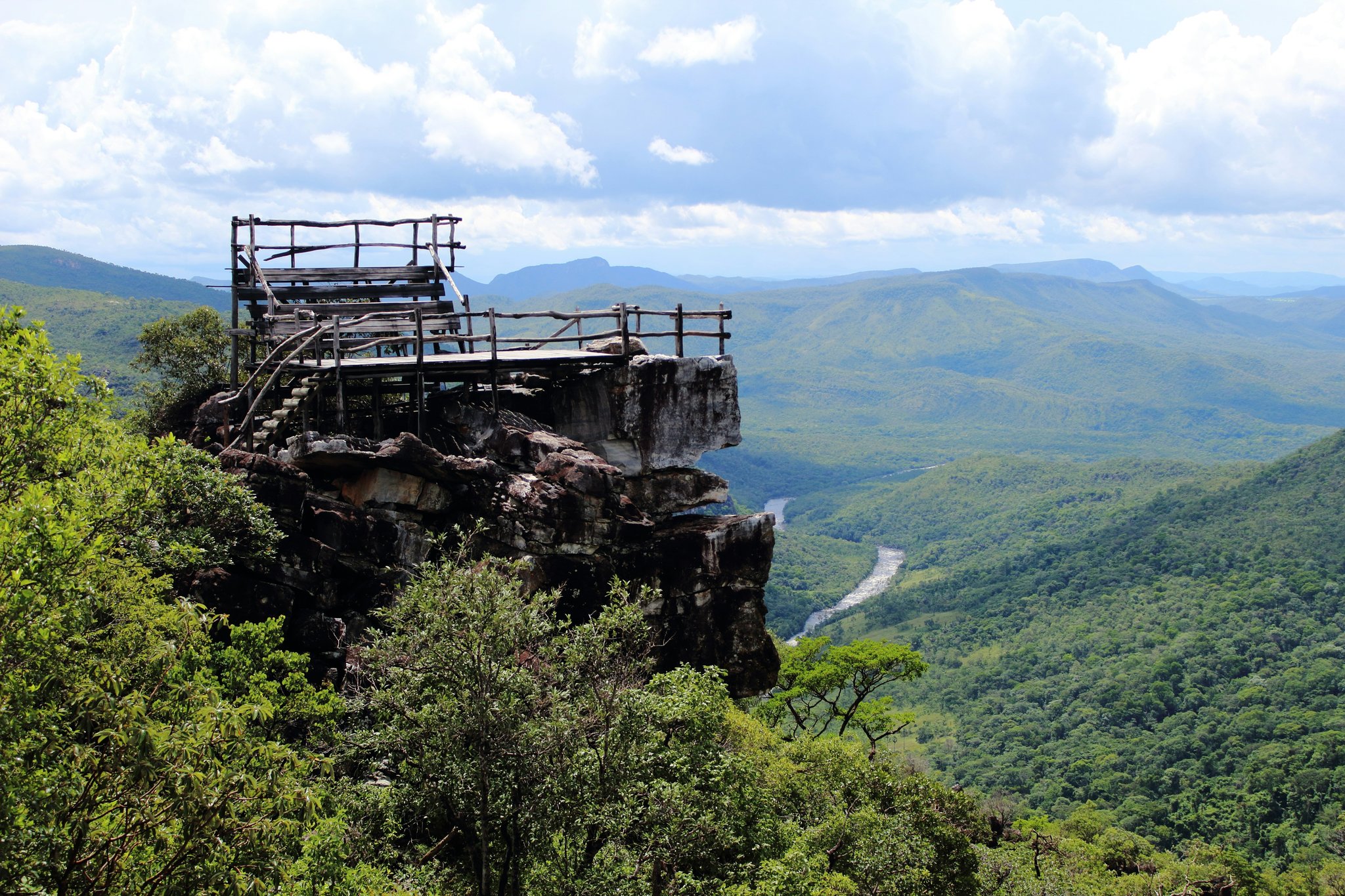 A rustic wooden viewpoint platform perched on a rocky cliff edge during trekking Chapada dos Veadeiros, overlooking a winding river and vast green cerrado valley below