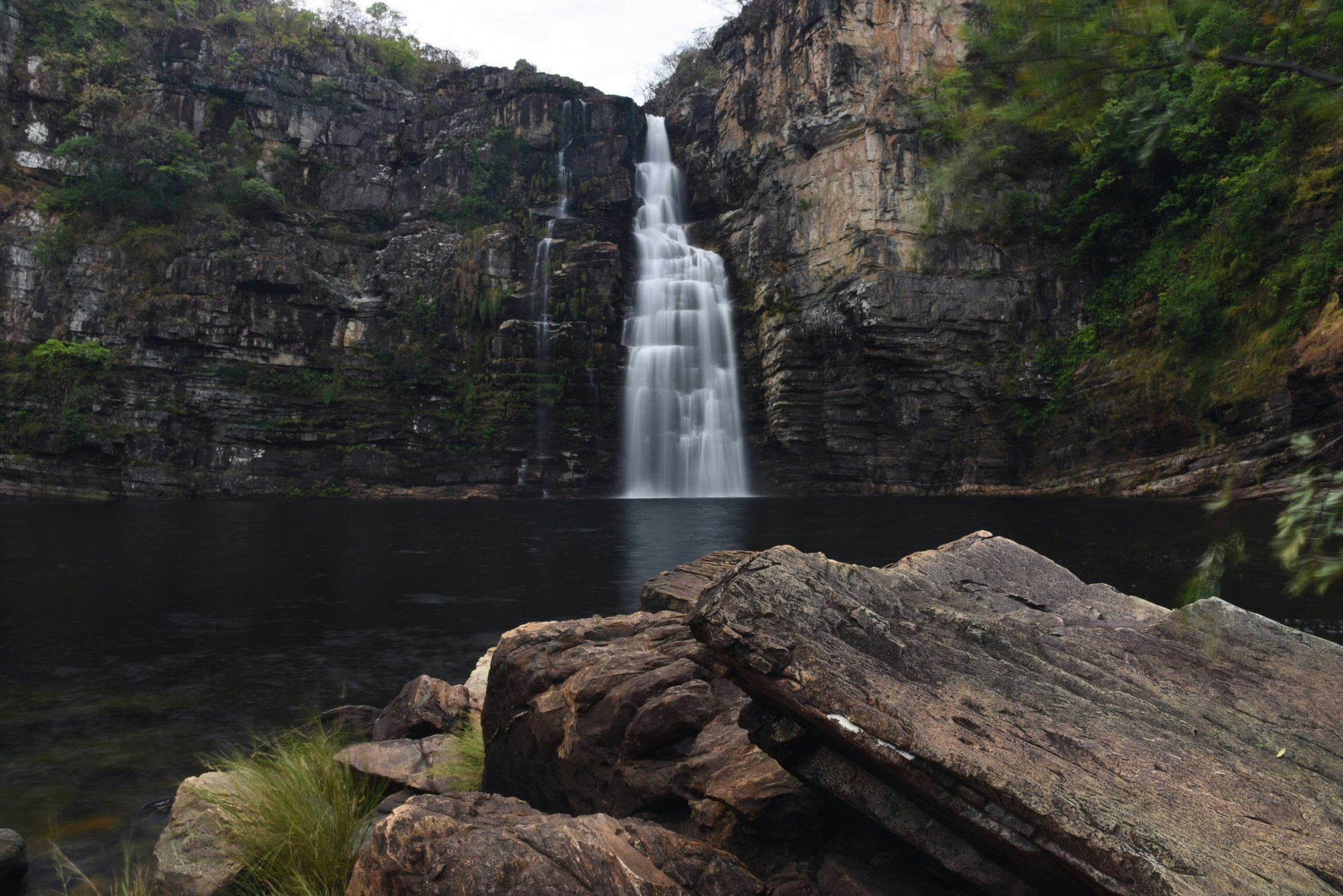 A tall multi-tiered waterfall cascades down a layered sandstone cliff into a dark still pool, framed by green cerrado vegetation during a trekking Chapada dos Veadeiros expedition