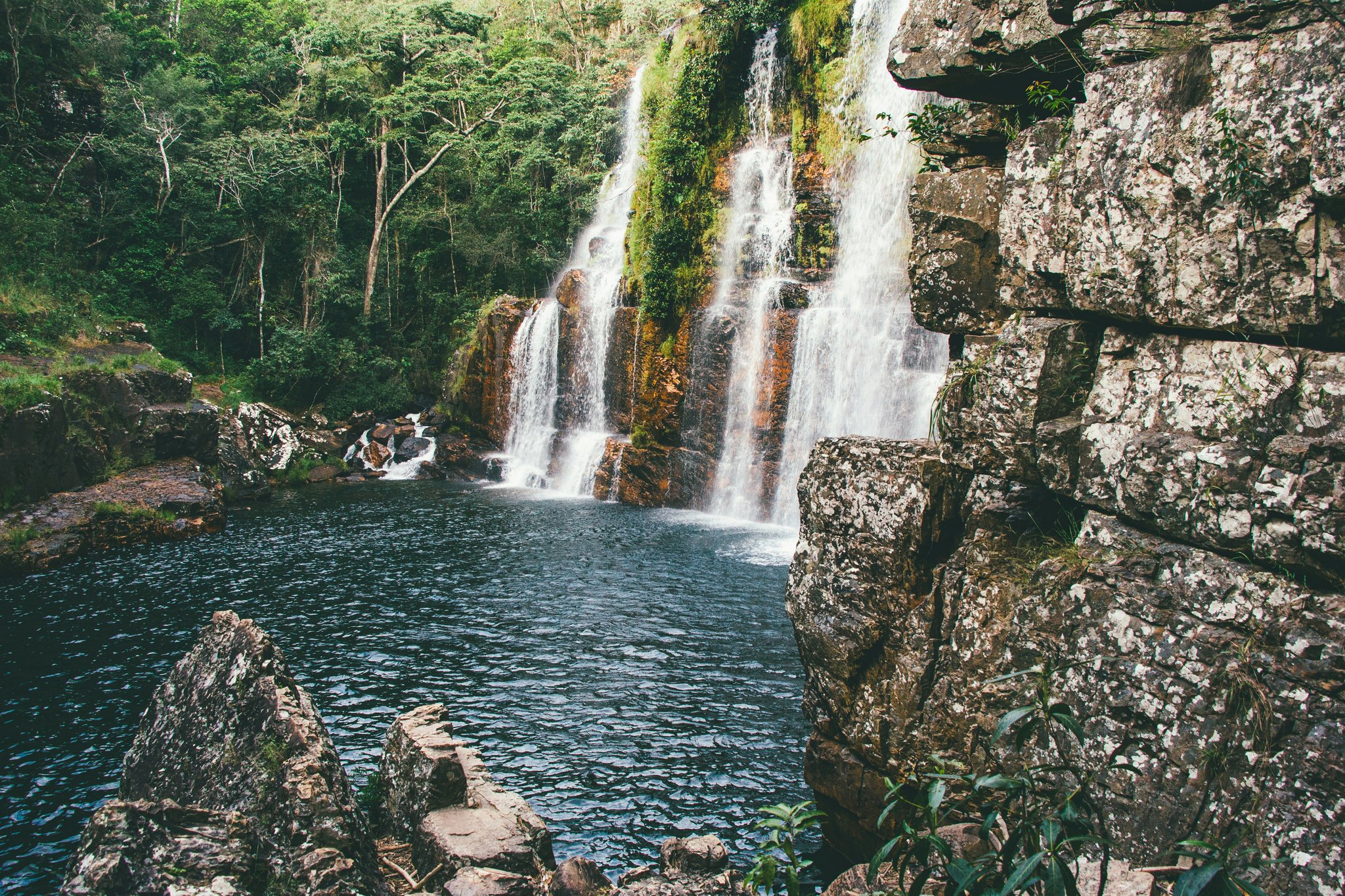 Multi-tiered waterfall cascading over moss-covered ochre rock faces into a dark natural pool, framed by dense Cerrado forest on a trekking Chapada dos Veadeiros route