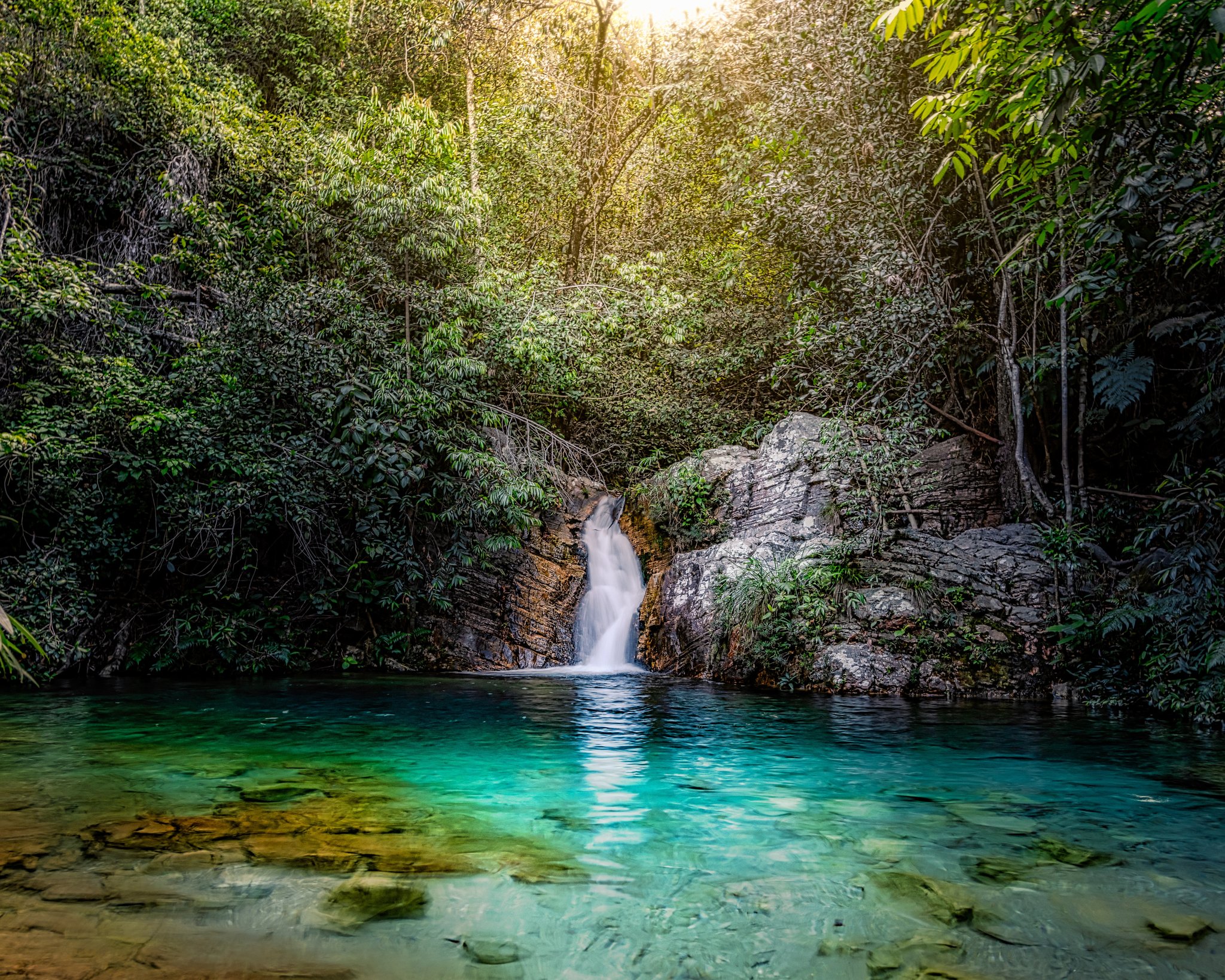 A silky waterfall cascades over layered rock into a crystal-clear turquoise natural pool surrounded by dense Cerrado rainforest canopy during a trekking Chapada dos Veadeiros adventure