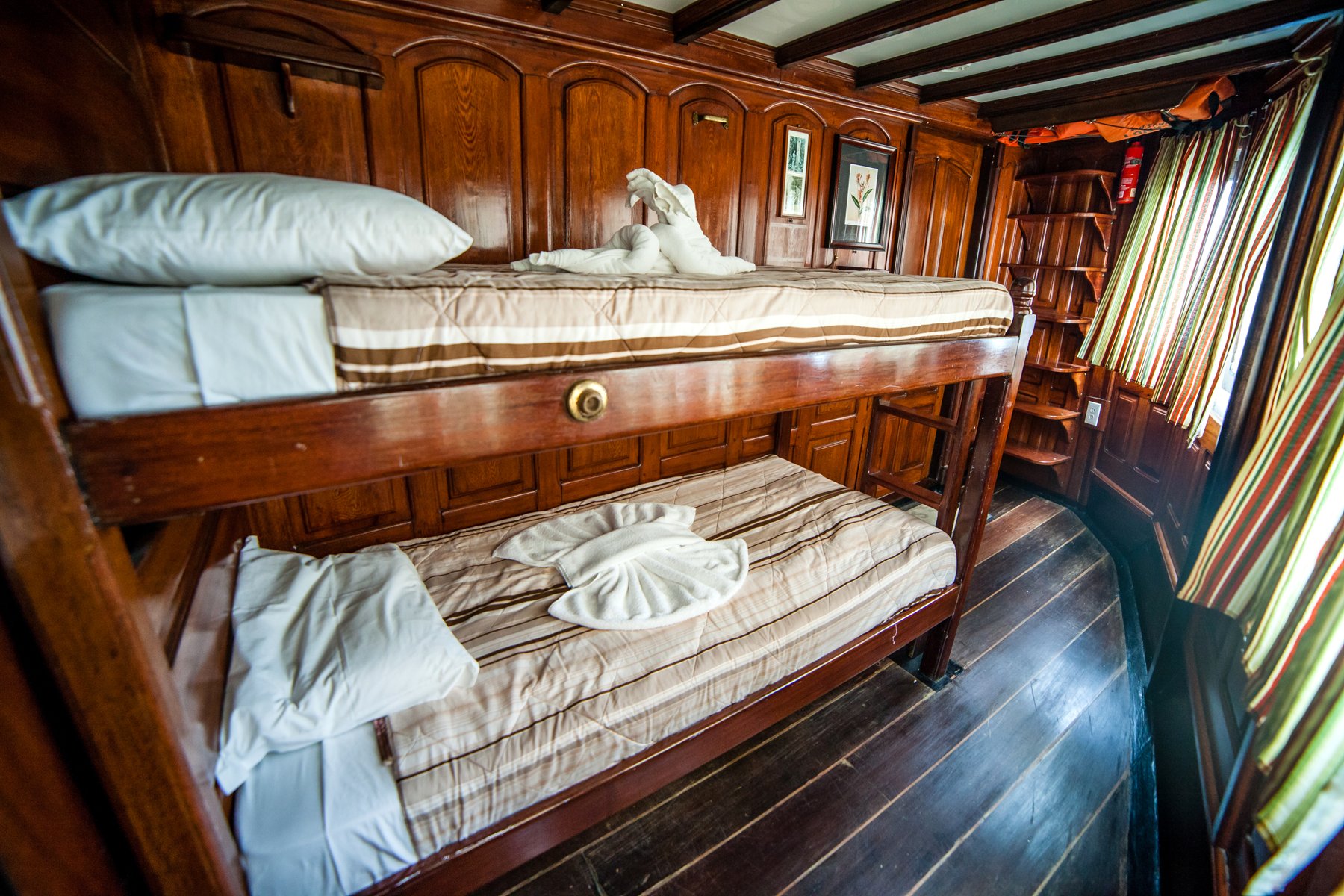 Polished wood-panelled bunk bed cabin aboard the Tucano Amazon river cruise, with striped bedding, folded towel sculptures and a small wooden bookshelf beside striped curtains
