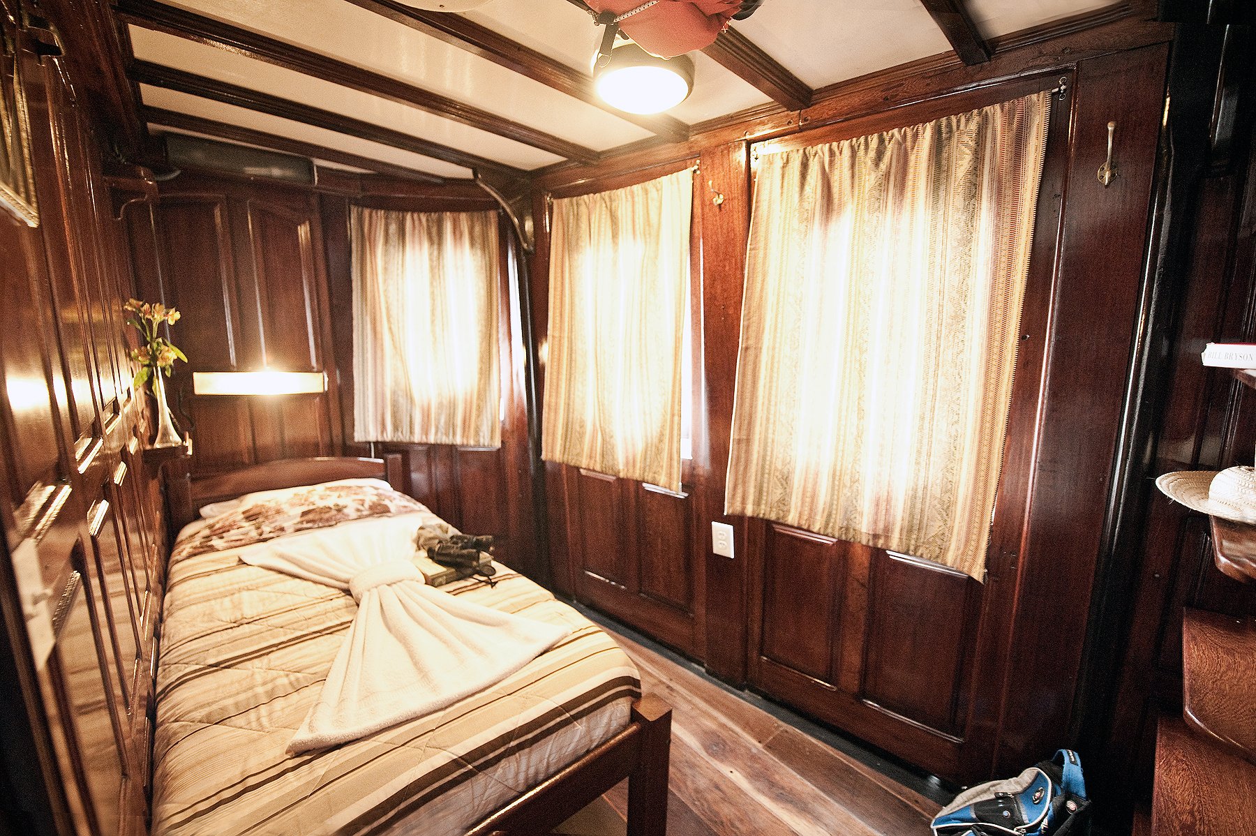 Cozy en-suite cabin aboard the Tucano Amazon river cruise, featuring dark varnished wood paneling, a made-up bed with striped linens, warm wall lighting, gold-toned curtained windows, and a small flower arrangement on the bedside shelf