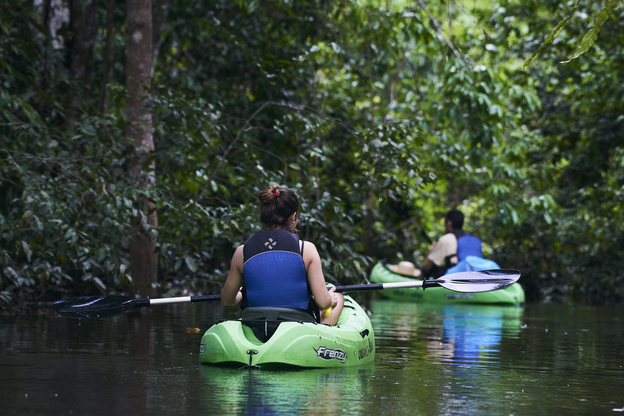 Green kayaks, mirror-still water, a tunnel of jungle closing in ahead. Photo: JUKAPAZ Two kayakers in green kayaks paddling through a narrow, tree-lined waterway on the Tucano Amazon river cruise, surrounded by lush flooded forest