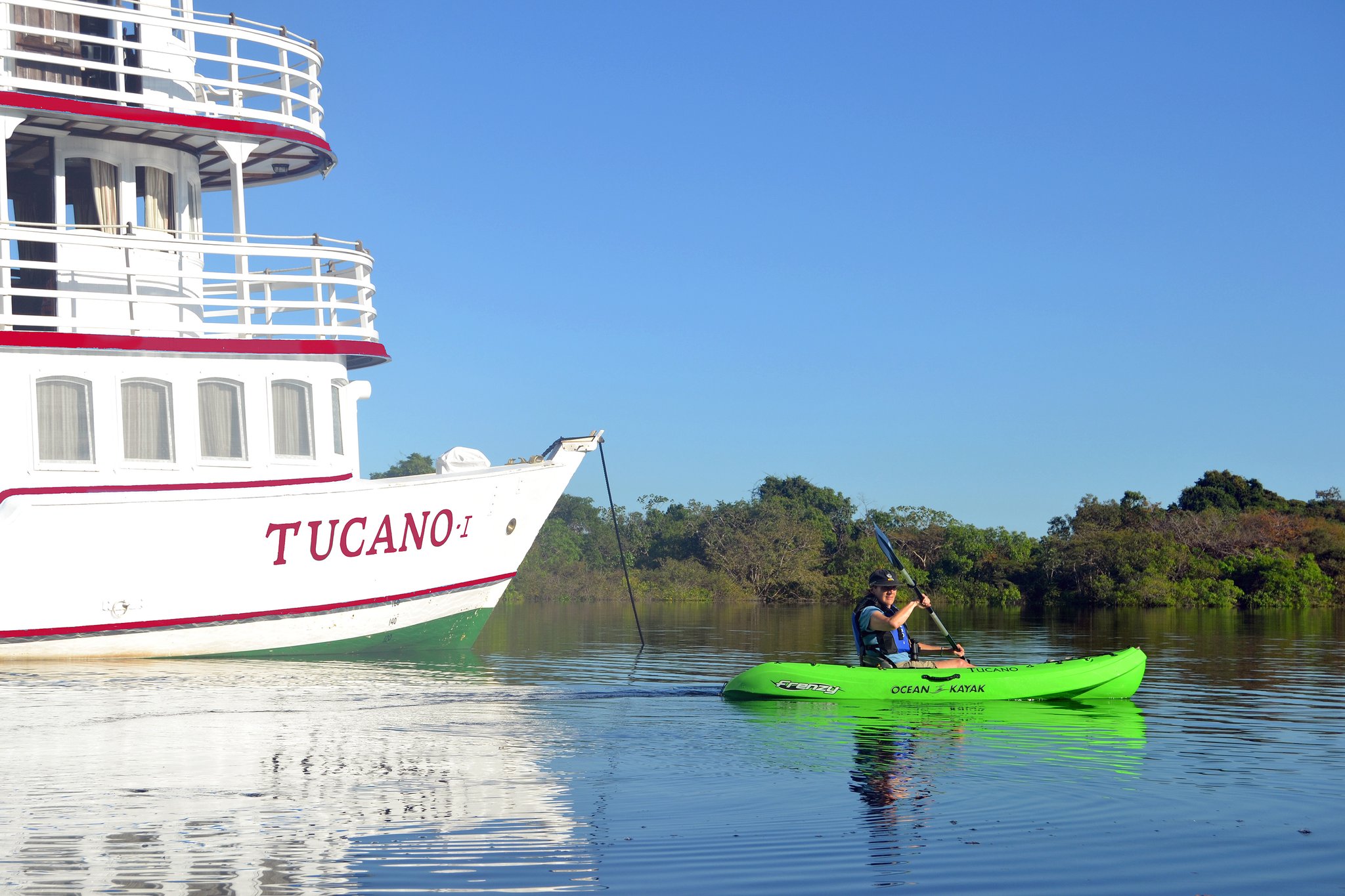 A traveler in a bright green kayak paddles alongside the bow of the M/Y Tucano on a calm river, with lush forest lining the far bank under a clear blue sky — one of the active excursions offered on the Tucano Amazon River Cruise