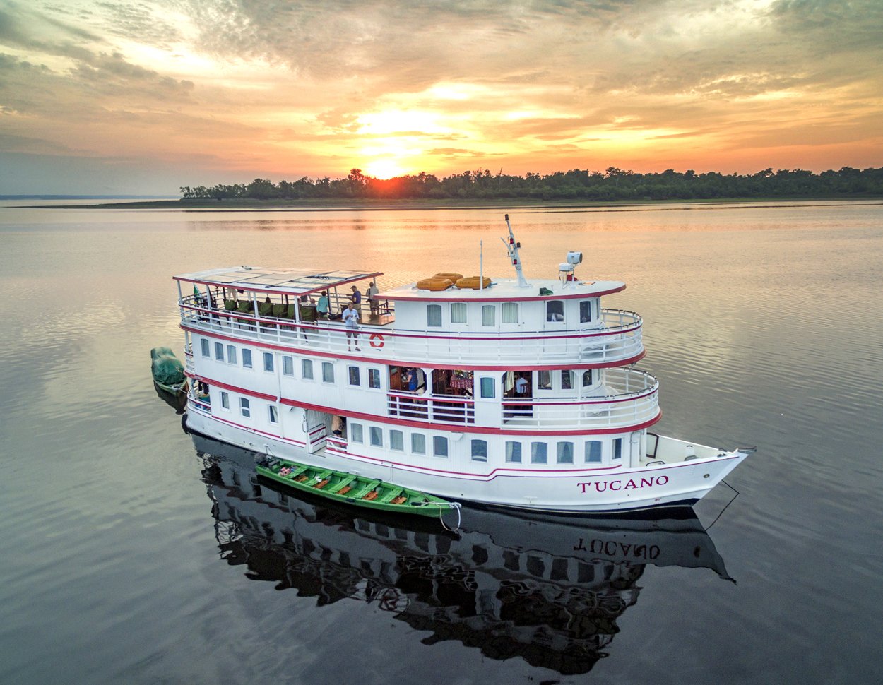 Sunset arrives quietly, and the Tucano sits perfectly still on the mirror-black river. The M/Y Tucano amazon river cruise vessel moored on glassy dark water at sunset, with guests on the upper observation deck and a green skiff alongside, forested horizon silhouetted against an orange sky