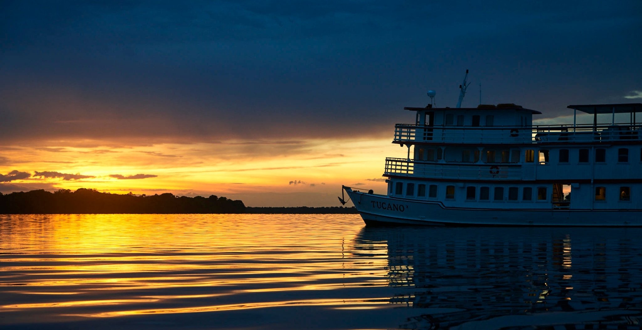 The M/Y Tucano amazon river cruise vessel moored at dusk, its white hull silhouetted against a blazing orange and gold sunset reflecting across a glassy river, with a dark forested shoreline on the horizon