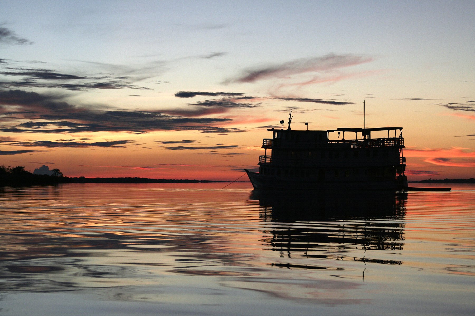 Fire in the sky, stillness on the water — the perfect close to a day deep in the Amazon. A multi-deck river vessel silhouetted against a blazing orange and crimson sunset reflected on glassy water during a tucano amazon river cruise