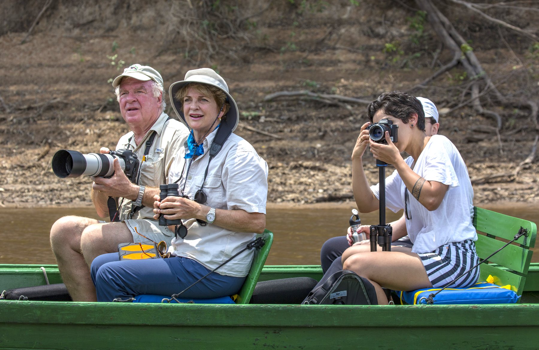 Cameras up, binoculars ready — every bend in the river brings a new discovery. Photo: Daniel Forster Three travelers with cameras and binoculars scan the riverbank from a green wooden boat on a tucano amazon river cruise excursion